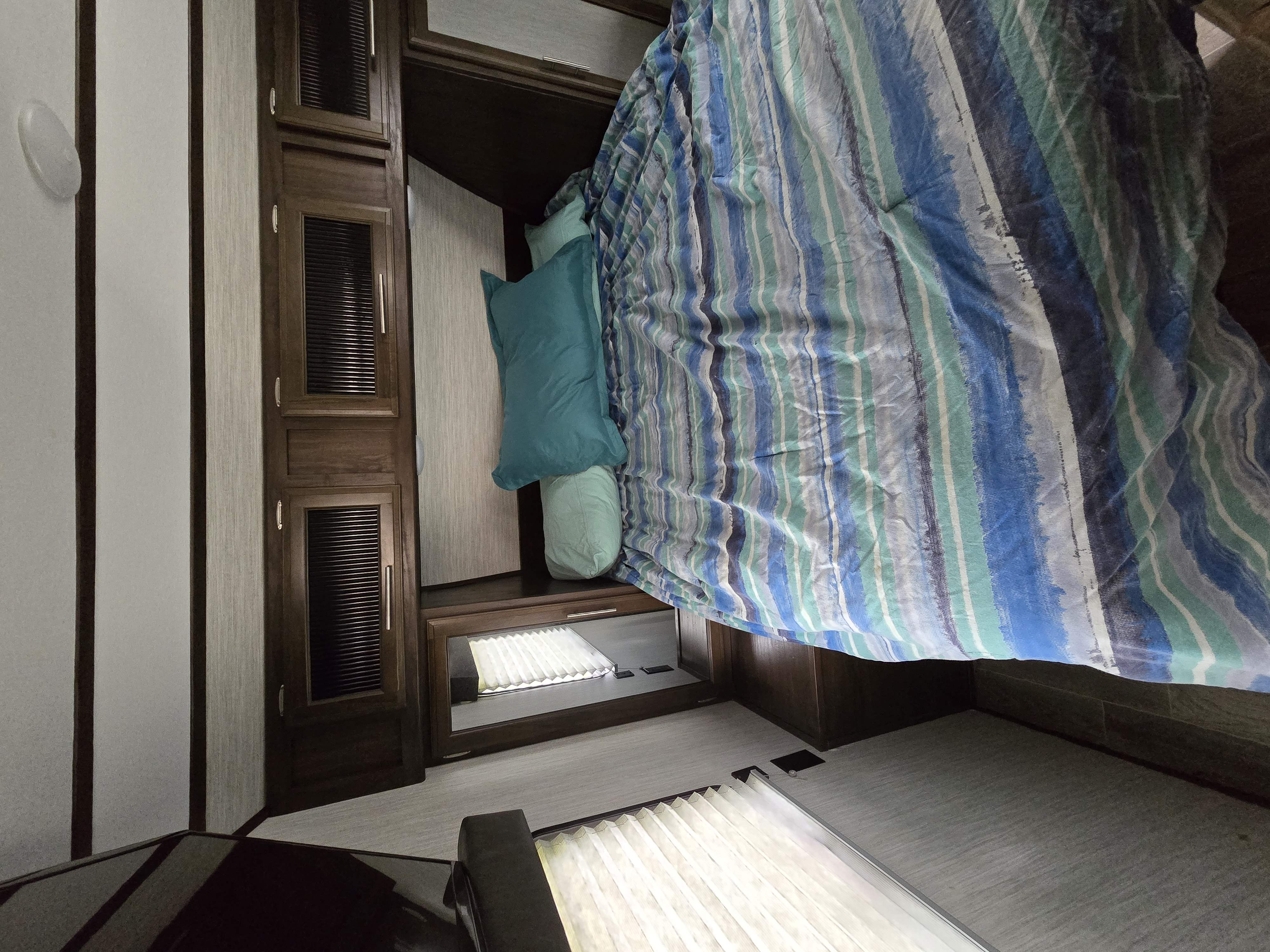 Cozy RV rental bedroom features a striped comforter, teal pillows, built-in dark wood cabinets, and lighted storage.. Forest River Cherokee Alpha Wolf 2019
