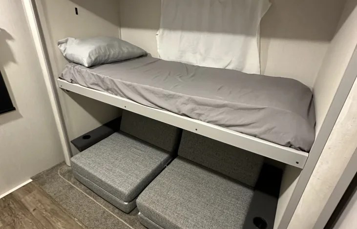 This compact RV rental features a cozy bunk bed with gray bedding above versatile gray seating, plus a small shelf with a cup holder.. Forest River Vibe 2022