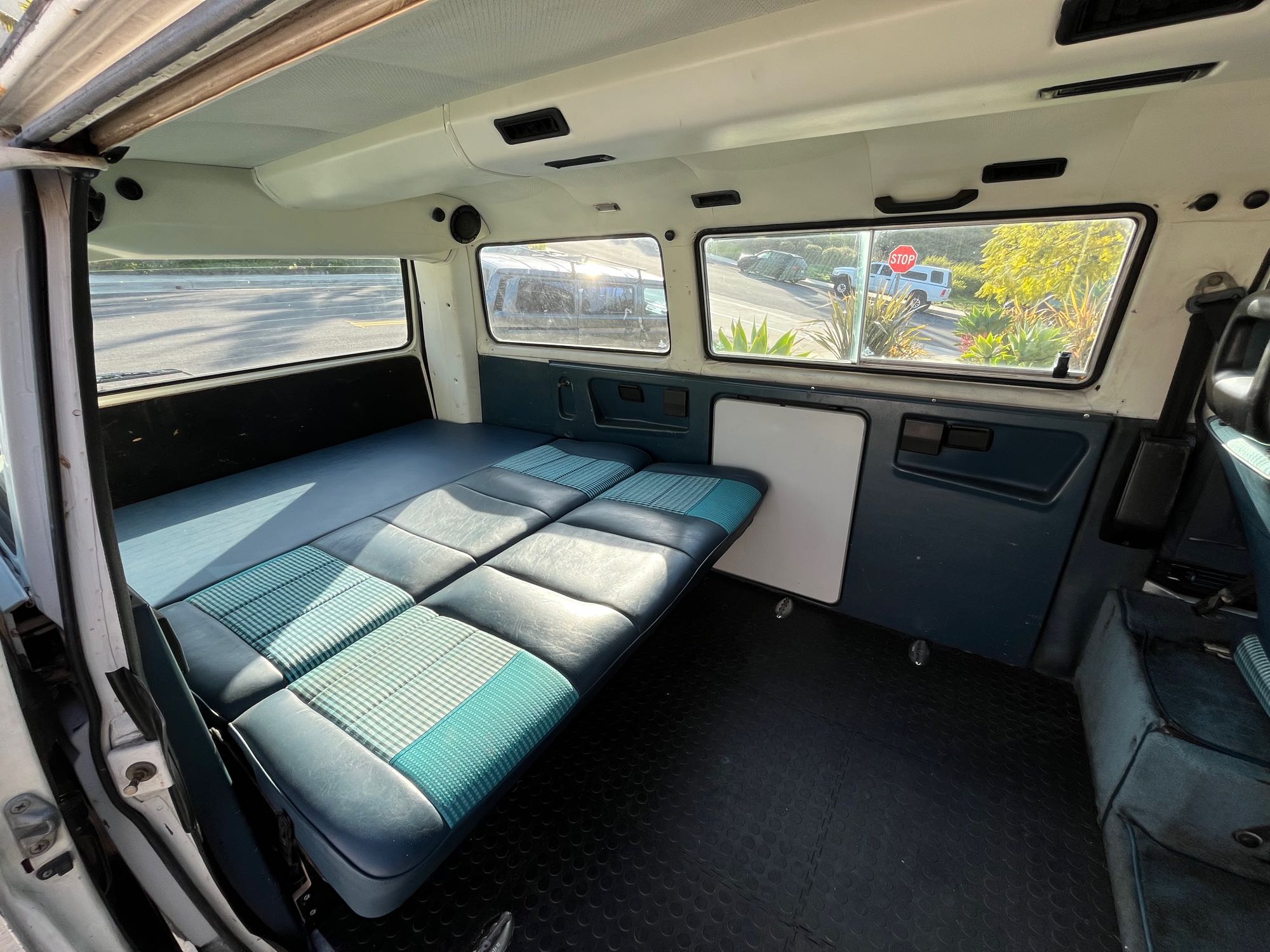 Comfortable RV rental interior: Blue/teal checkered bed, dark floor, and windows showing the street.. Volkswagen Vanagon 1987