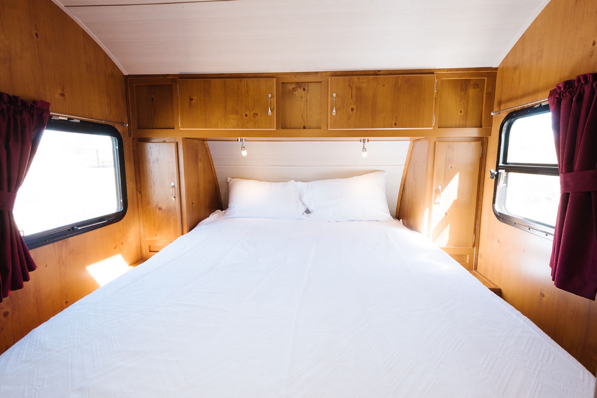 Cozy RV rental bedroom with a white bed, warm wooden interior, and bright windows with red curtains.. Gulf Stream Vintage Cruiser 2018