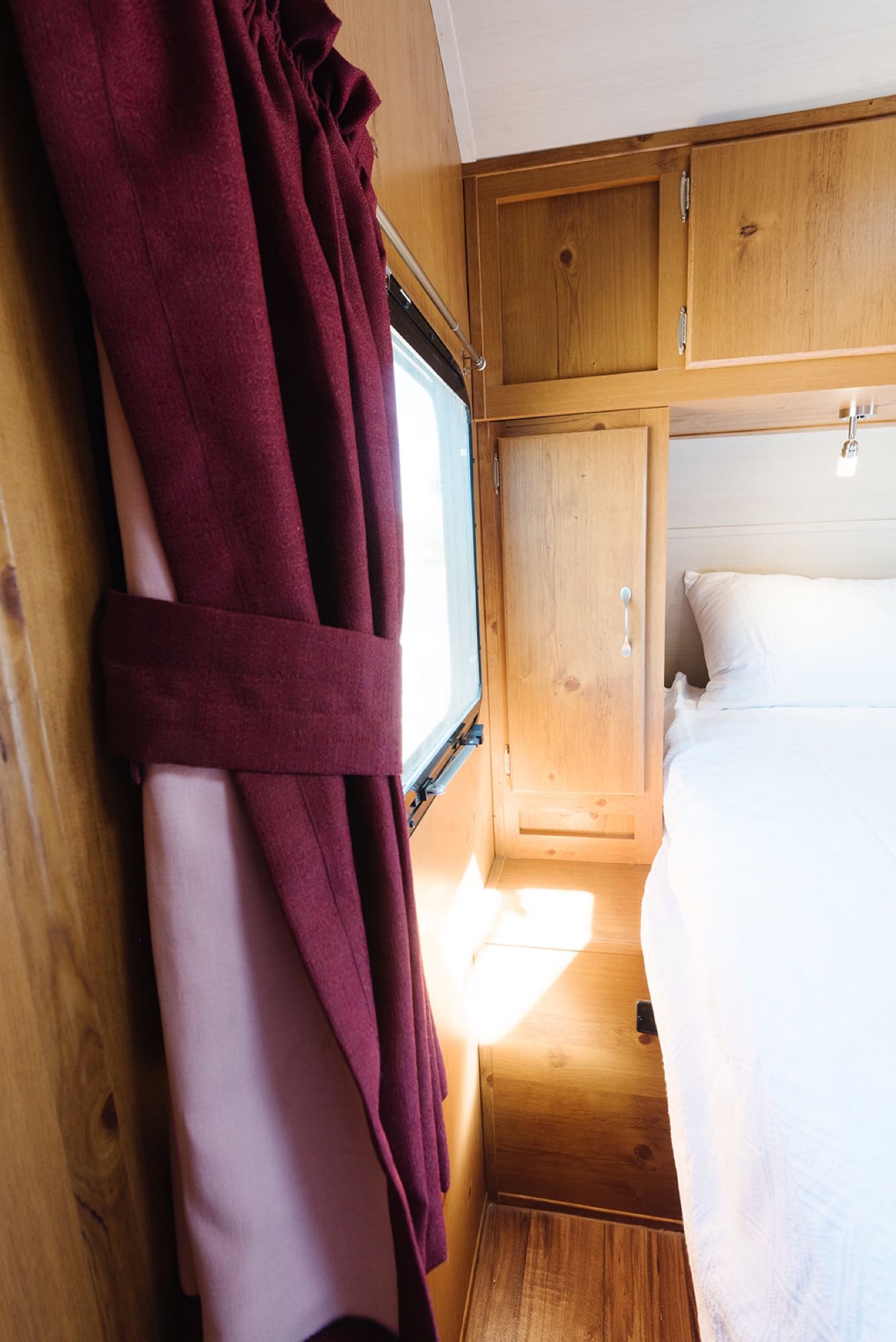 Bright RV rental interior: wooden walls, cozy bed with white linens, and a sunlit window with burgundy curtains.. Gulf Stream Vintage Cruiser 2018