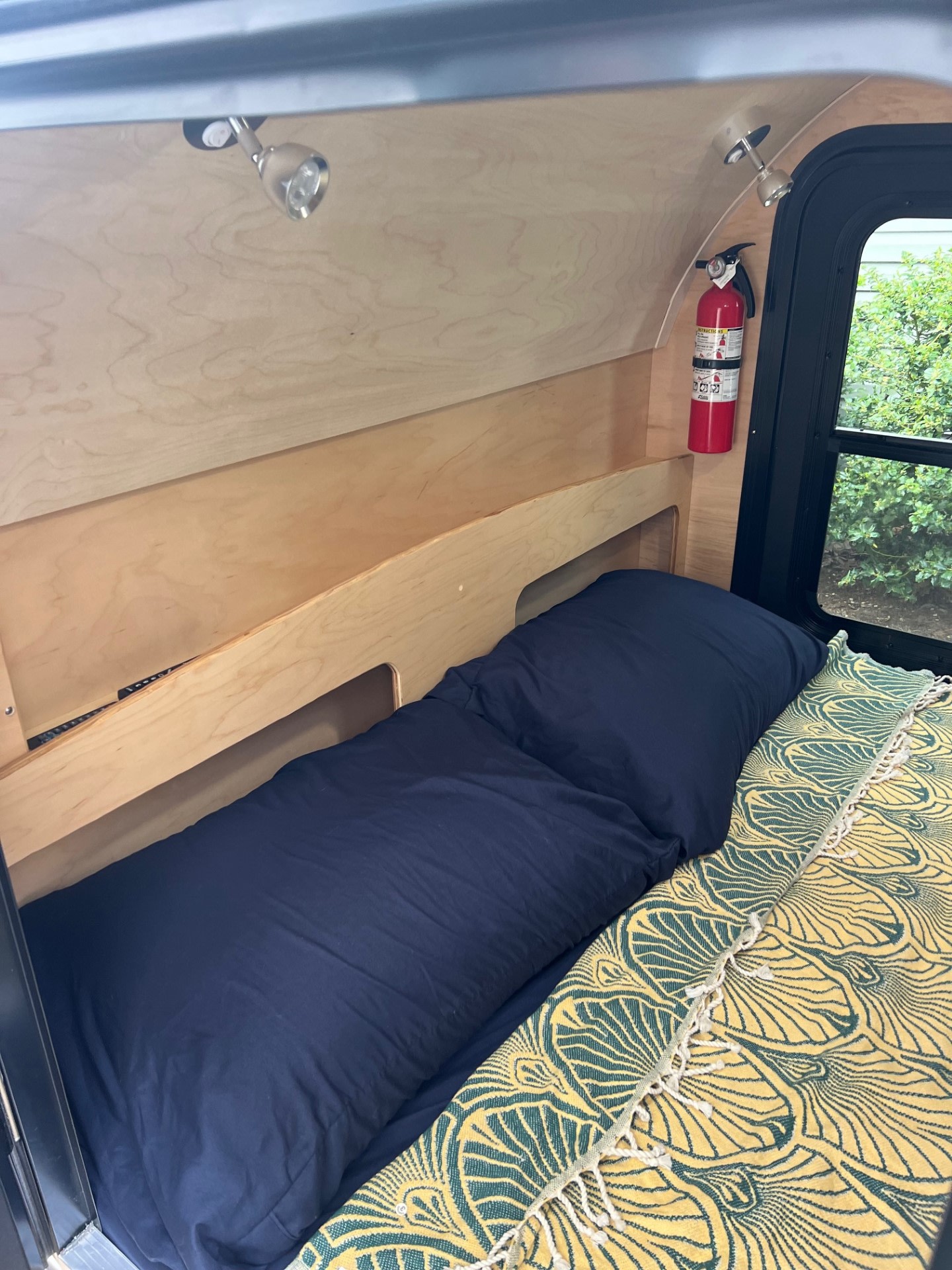 Inside a cozy RV rental: bed with blue linens & patterned throw, built-in shelf, fire extinguisher, window.. UkanCamp UkanCamp Trailer 2023