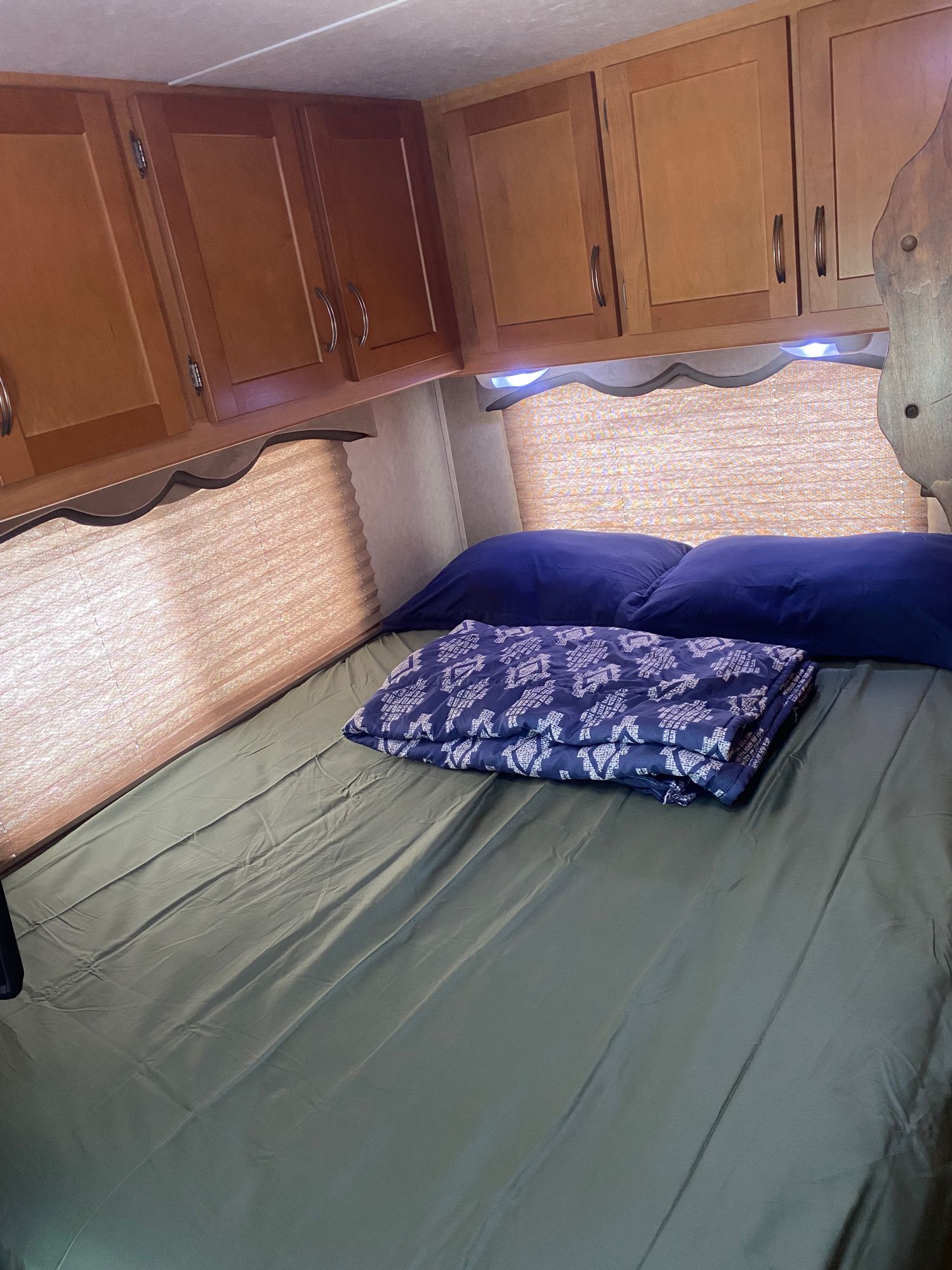This cozy RV rental bedroom features a neatly made bed with blue pillows and a blanket, under wooden cabinets and soft lighting.. Forest River Sunseeker 2009