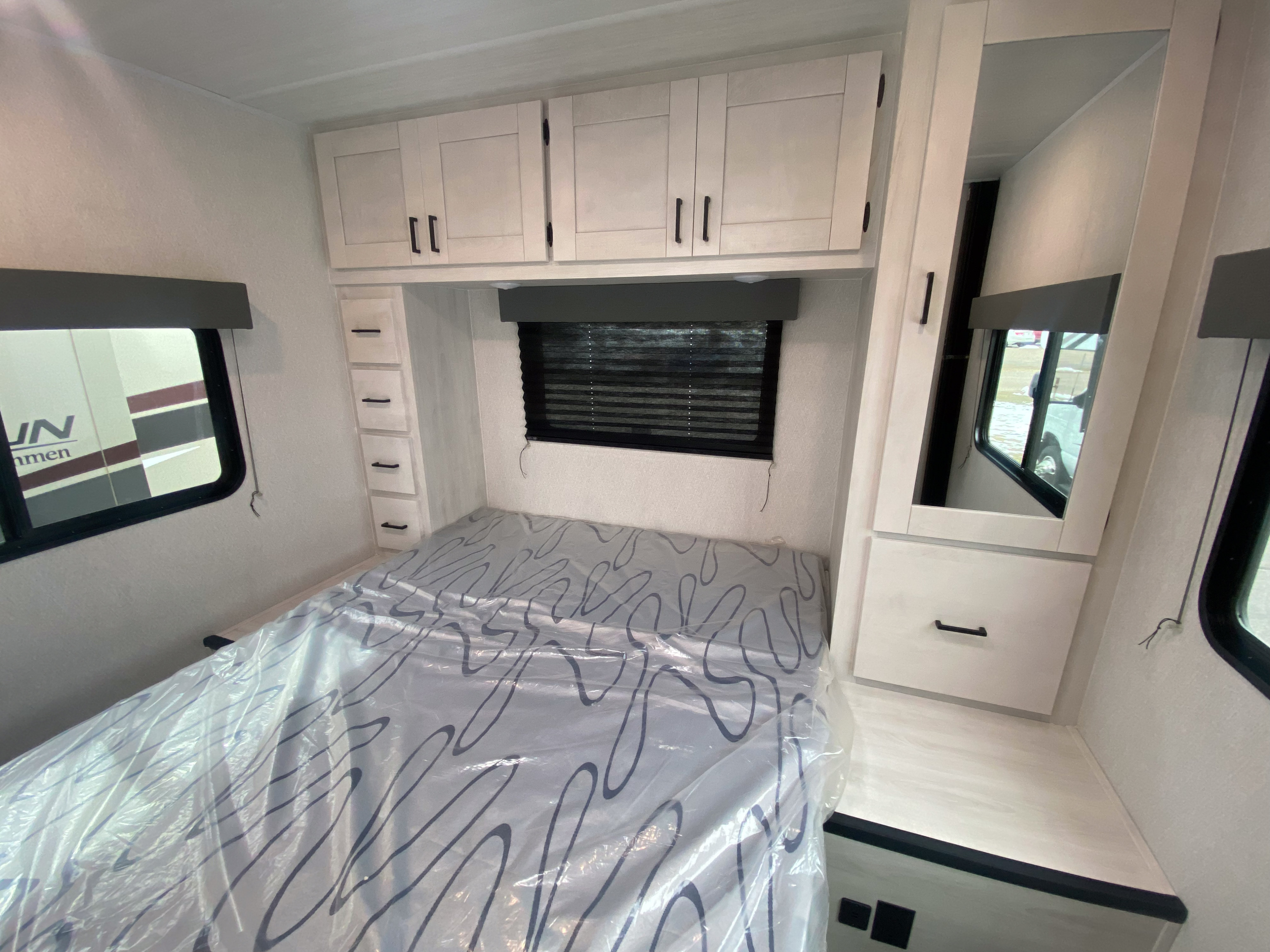Spacious RV rental bedroom with a new, plastic-covered bed, white overhead cabinets, and a built-in mirrored wardrobe.. East to West Entrada 2025