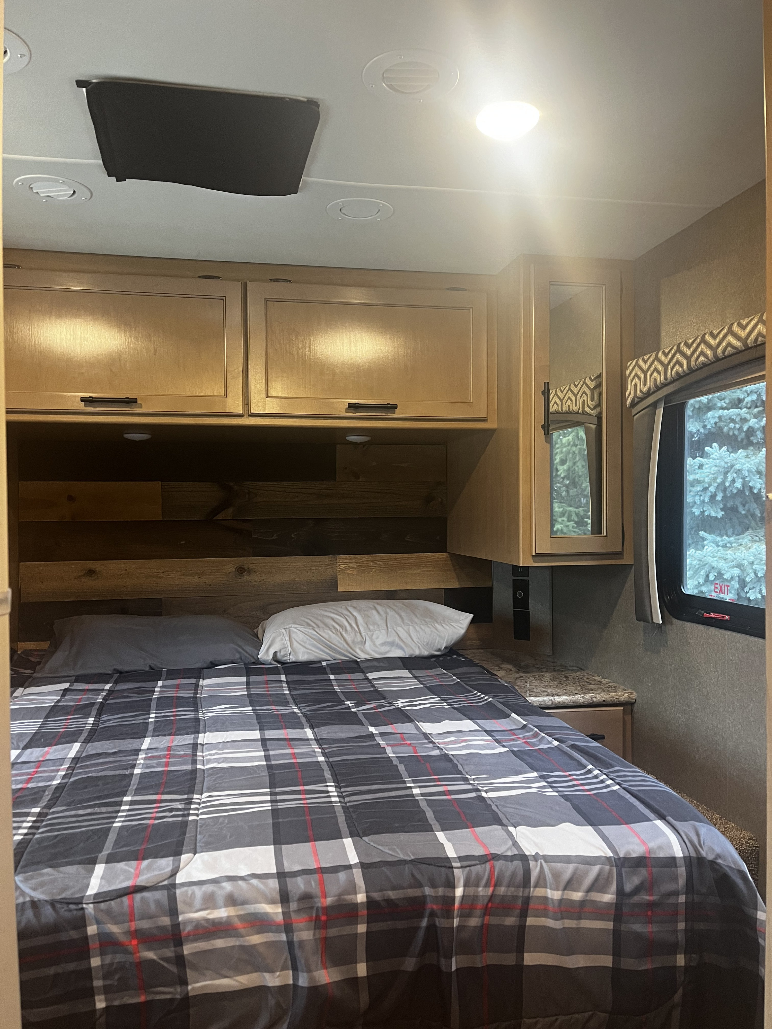 Cozy RV rental bedroom featuring a plaid comforter, wood headboard, overhead cabinets, and a window with a treed view.. Thor Chateau 2021