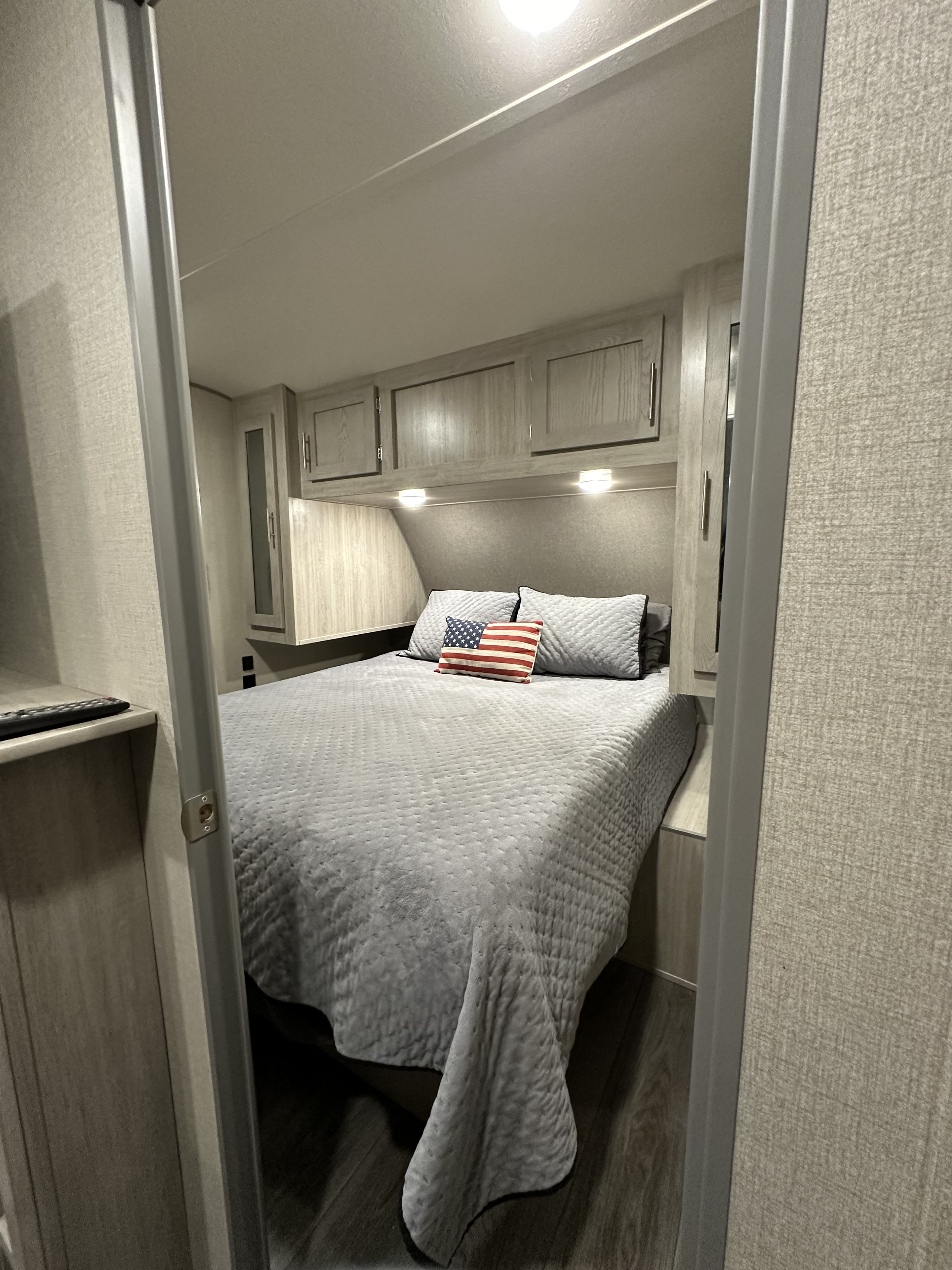 Cozy RV rental bedroom with light grey bedding, an American flag pillow, and compact overhead cabinets.. Coachmen Catalina Legacy Edition 2022