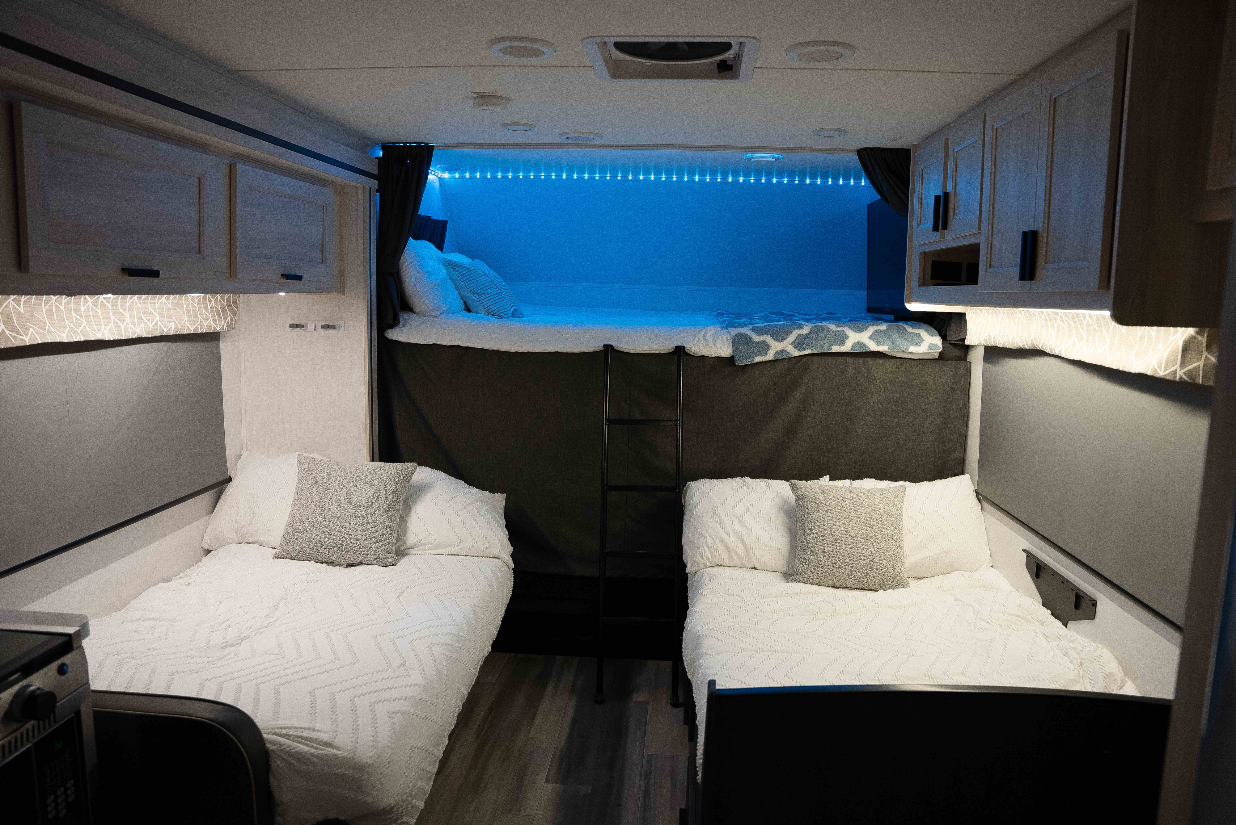 This RV rental features a modern interior with two lower beds, an upper bunk with blue accent lighting, and overhead cabinets.. Forest River Sunseeker LE 2023