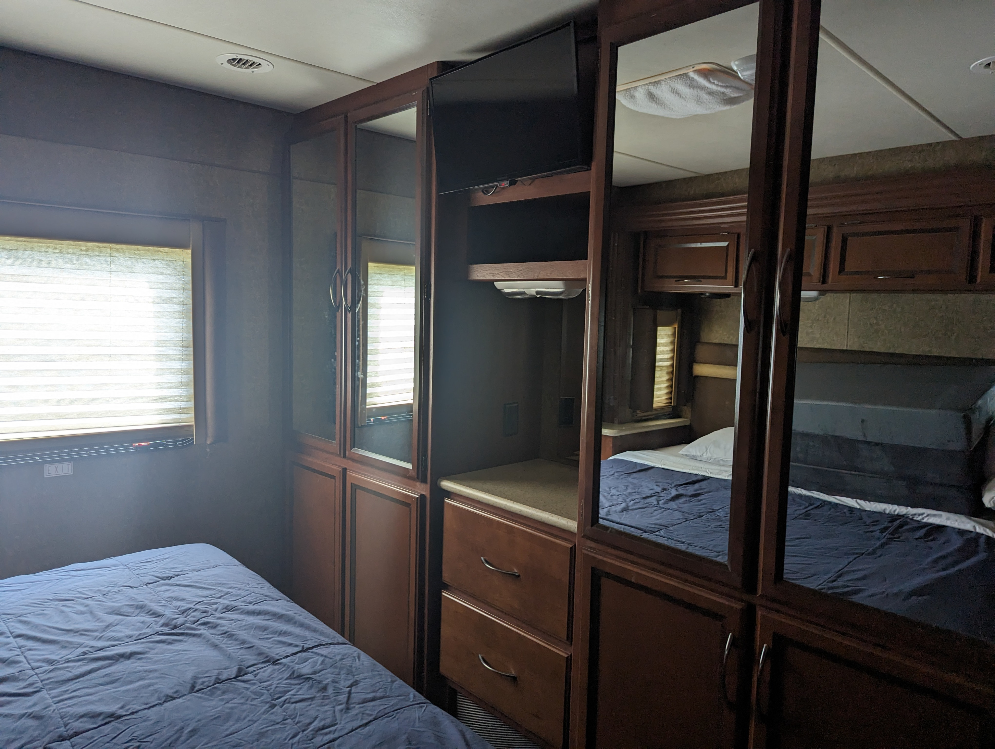 Spacious RV rental bedroom with a comfy blue bed, window, and wood cabinetry featuring mirrors, TV, and storage.. Thor Other 2014