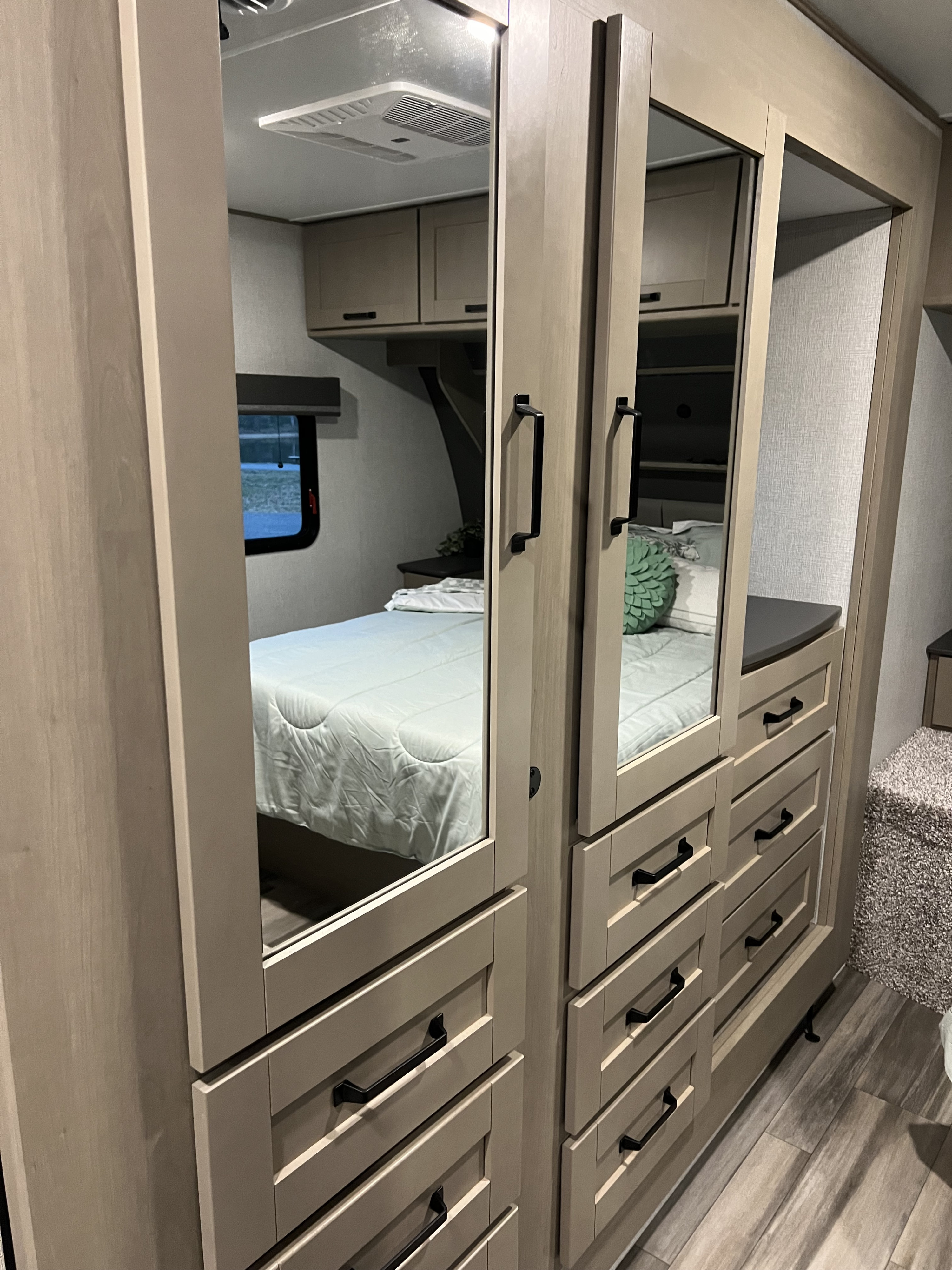 This inviting RV rental bedroom features a mirrored wardrobe with drawers, reflecting the cozy bed and a nature view.. Grand Design Reflection 2024