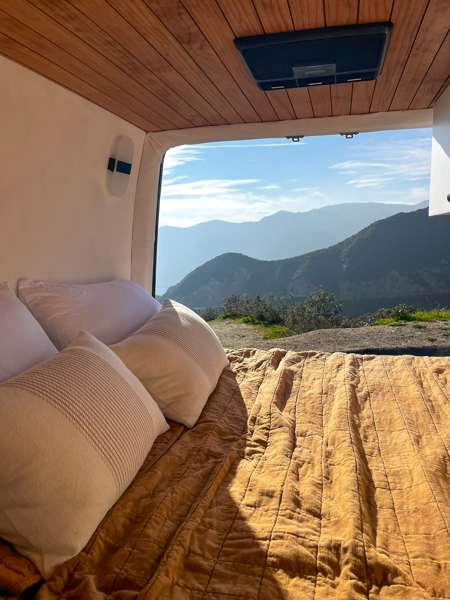 Inside an RV rental, a comfy bed with a quilted blanket overlooks beautiful layered mountains under a blue sky.. Mercedes Sprinter 2023