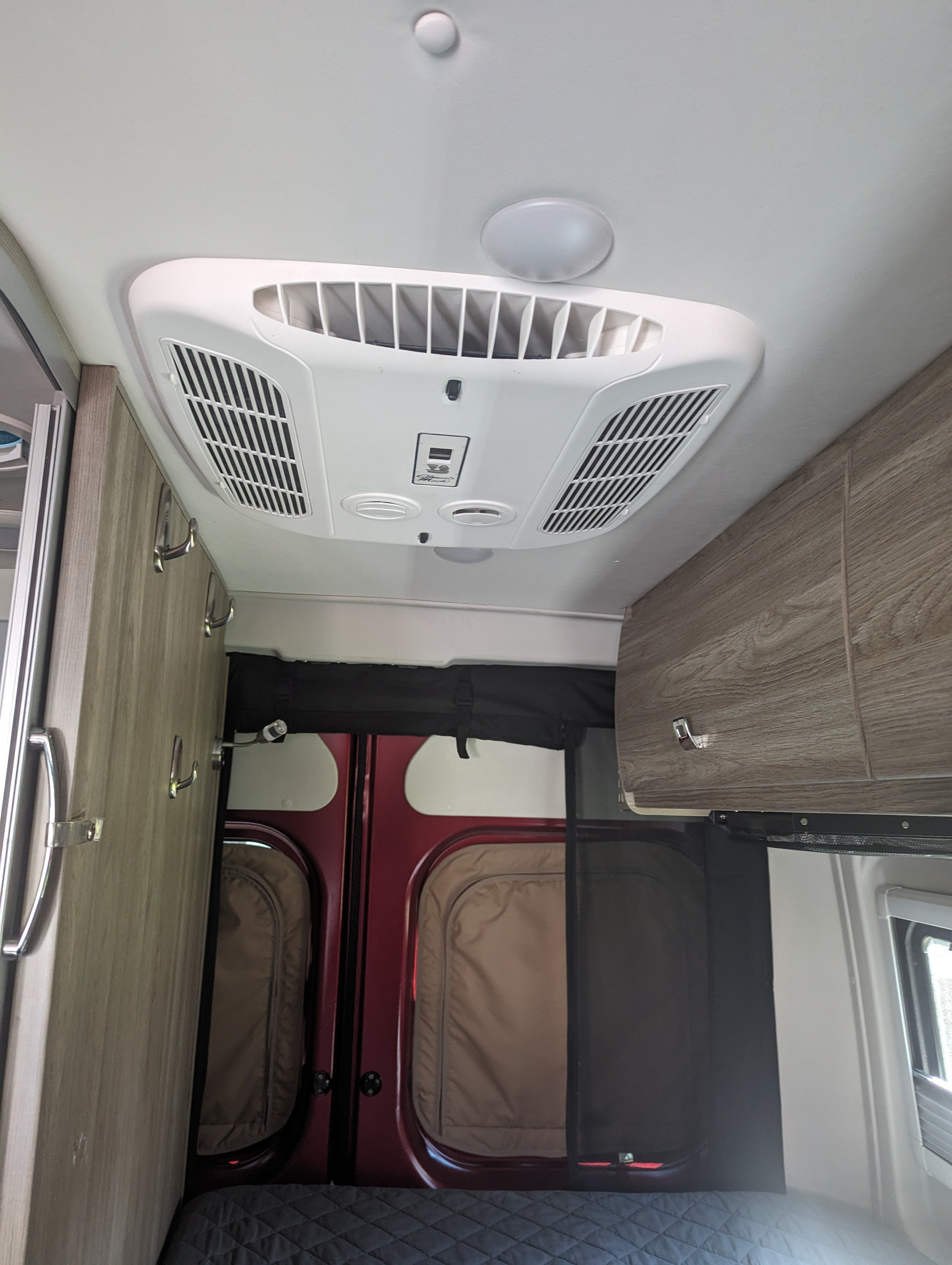 Overhead view in an rv rental showing a Dometic AC unit, ceiling lights, side cabinets with hooks, and rear doors with screens.. Winnebago Travato 2023