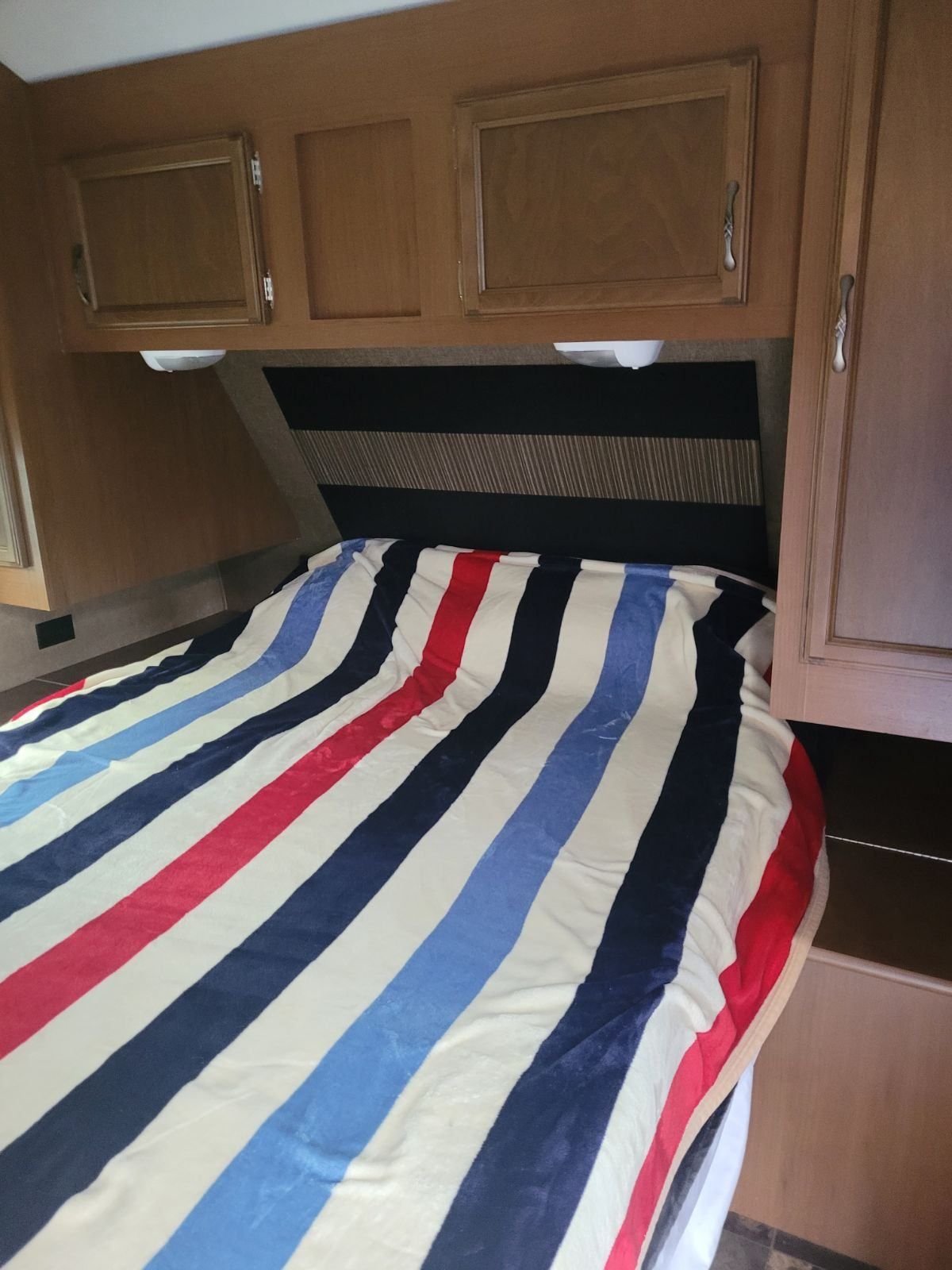 RV rental bedroom features a bed with a striped blanket, wooden storage cabinets, and overhead reading lights.. Starcraft Autumn Ridge 2014