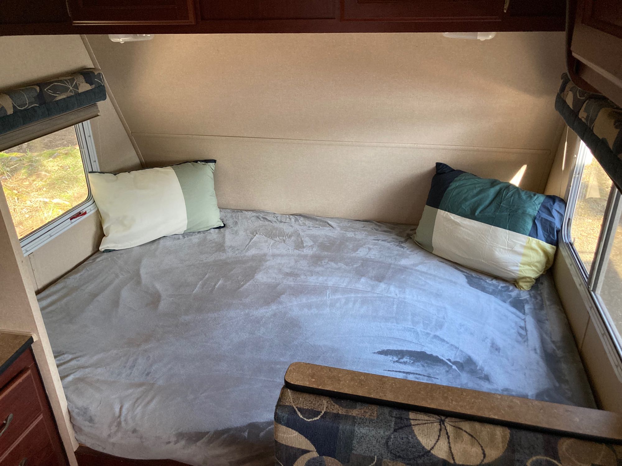 An RV rental interior showing a large bed with grey bedding and colorful pillows, flanked by bright windows.. Takena Takena Trailer 2010