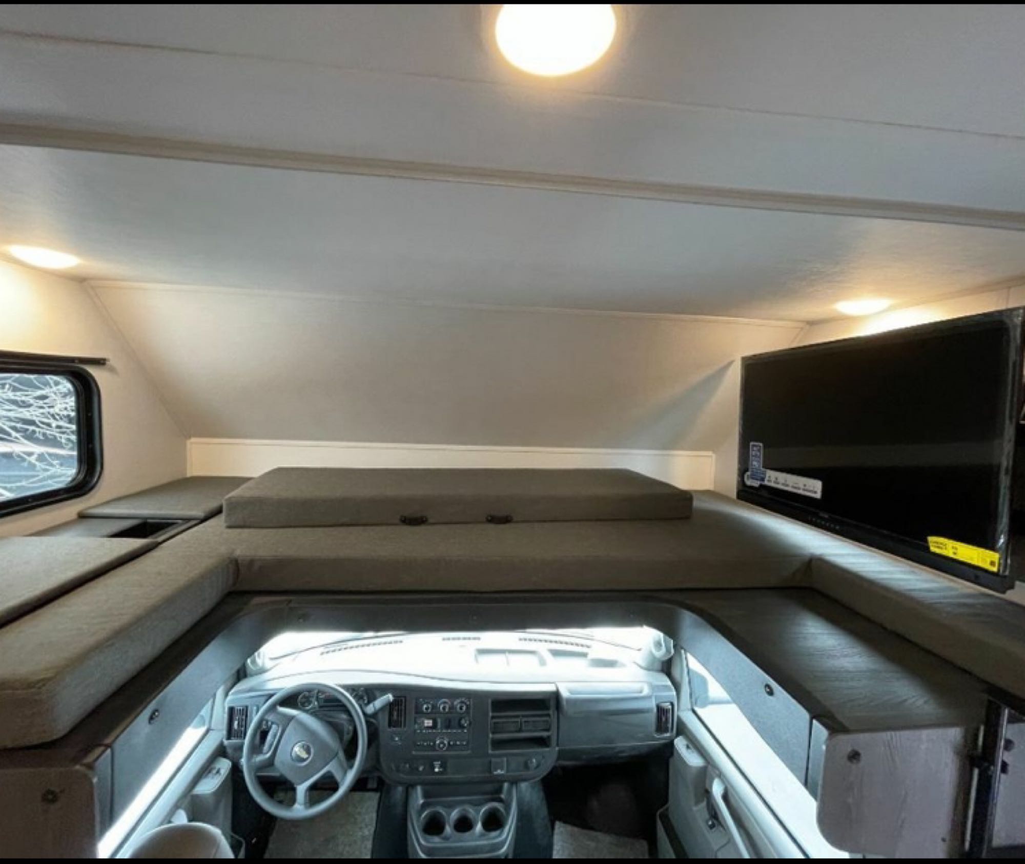 RV rental interior: cozy over-cab bunk with grey cushions, TV, and driver's cab below. Perfect for comfortable road trips.. Forester LE 2023
