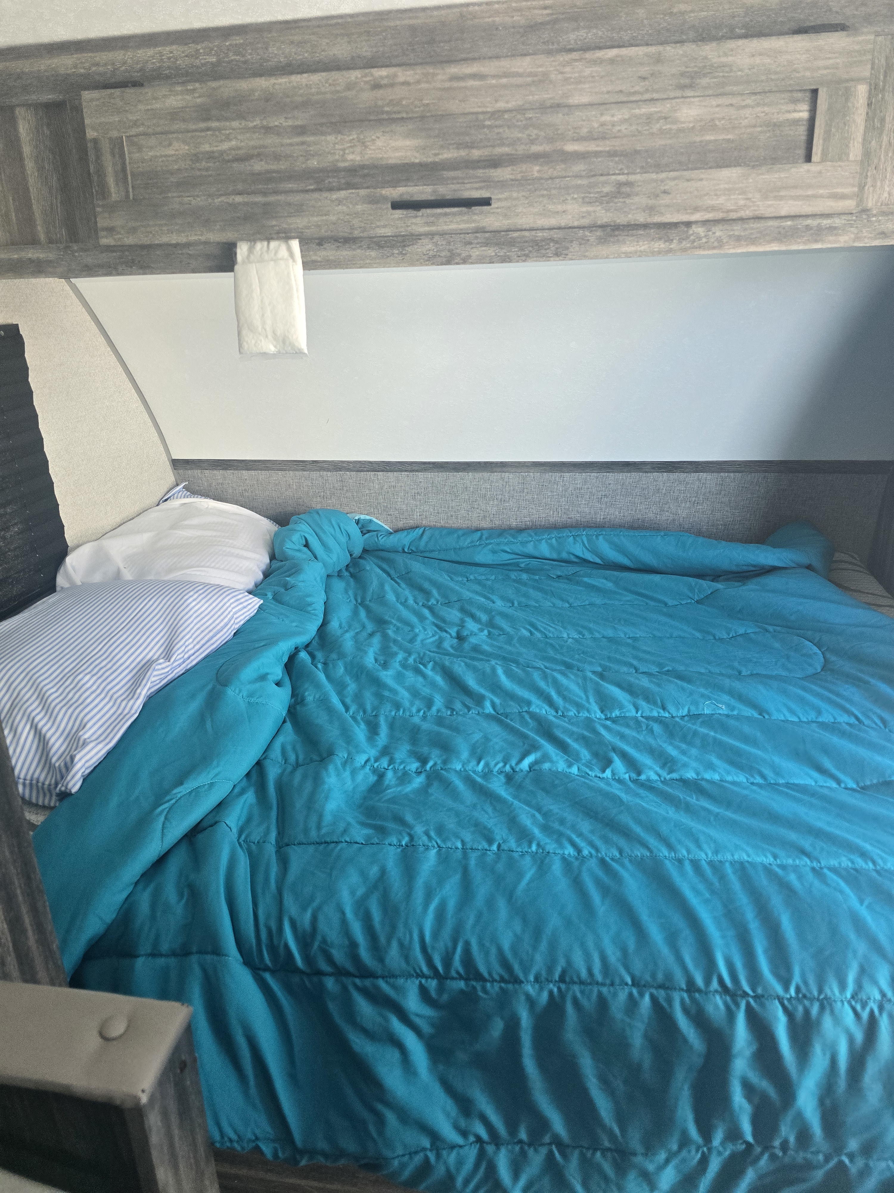 Cozy bed inside an RV rental with a bright turquoise comforter, striped and white pillows, and wood-look cabinets overhead.. Forest River Wildwood FSX 2021