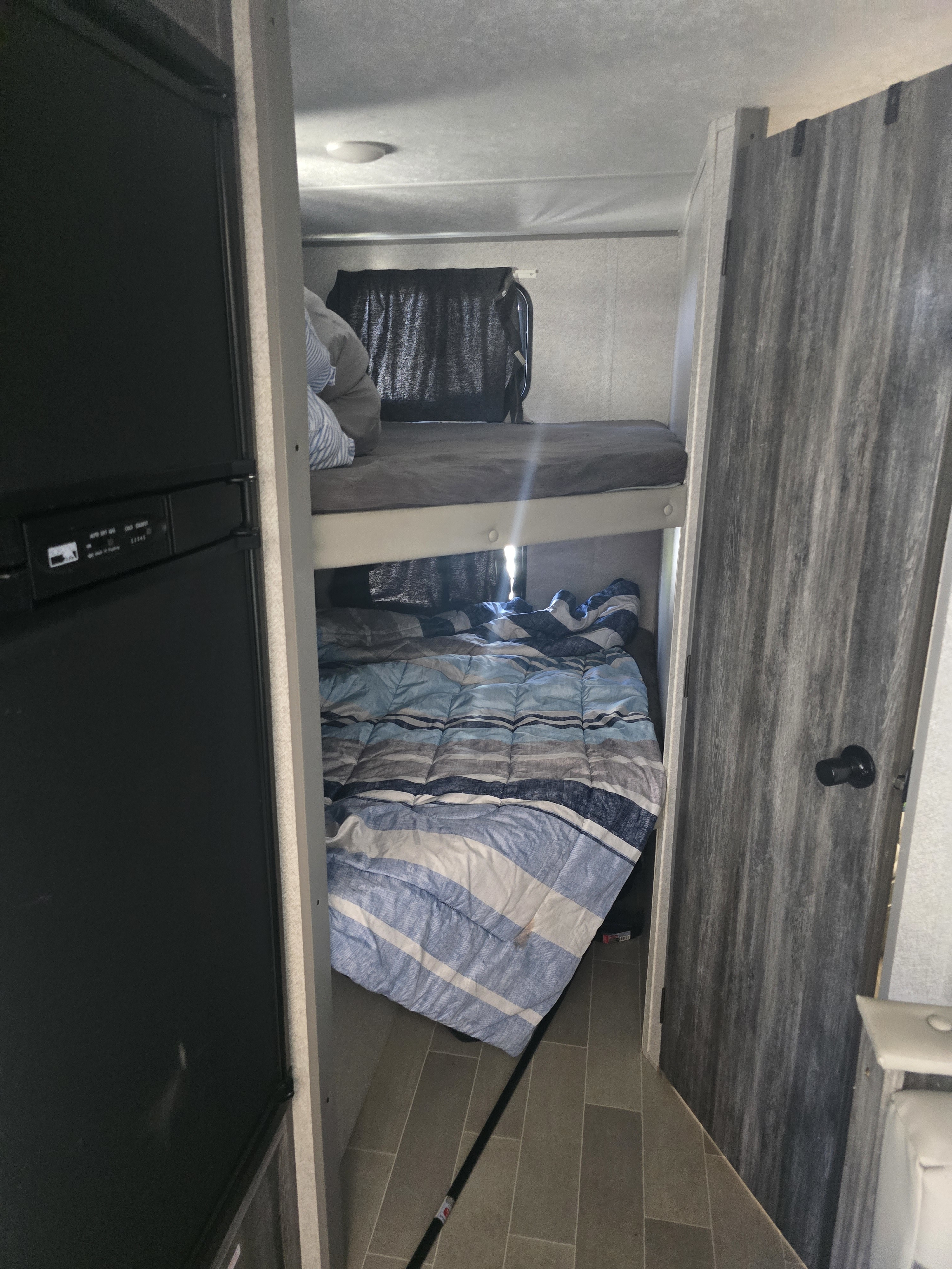 Compact RV rental interior featuring cozy bunk beds with striped bedding, a fridge, and a door, designed for efficient sleeping in a small space.. Forest River Wildwood FSX 2021