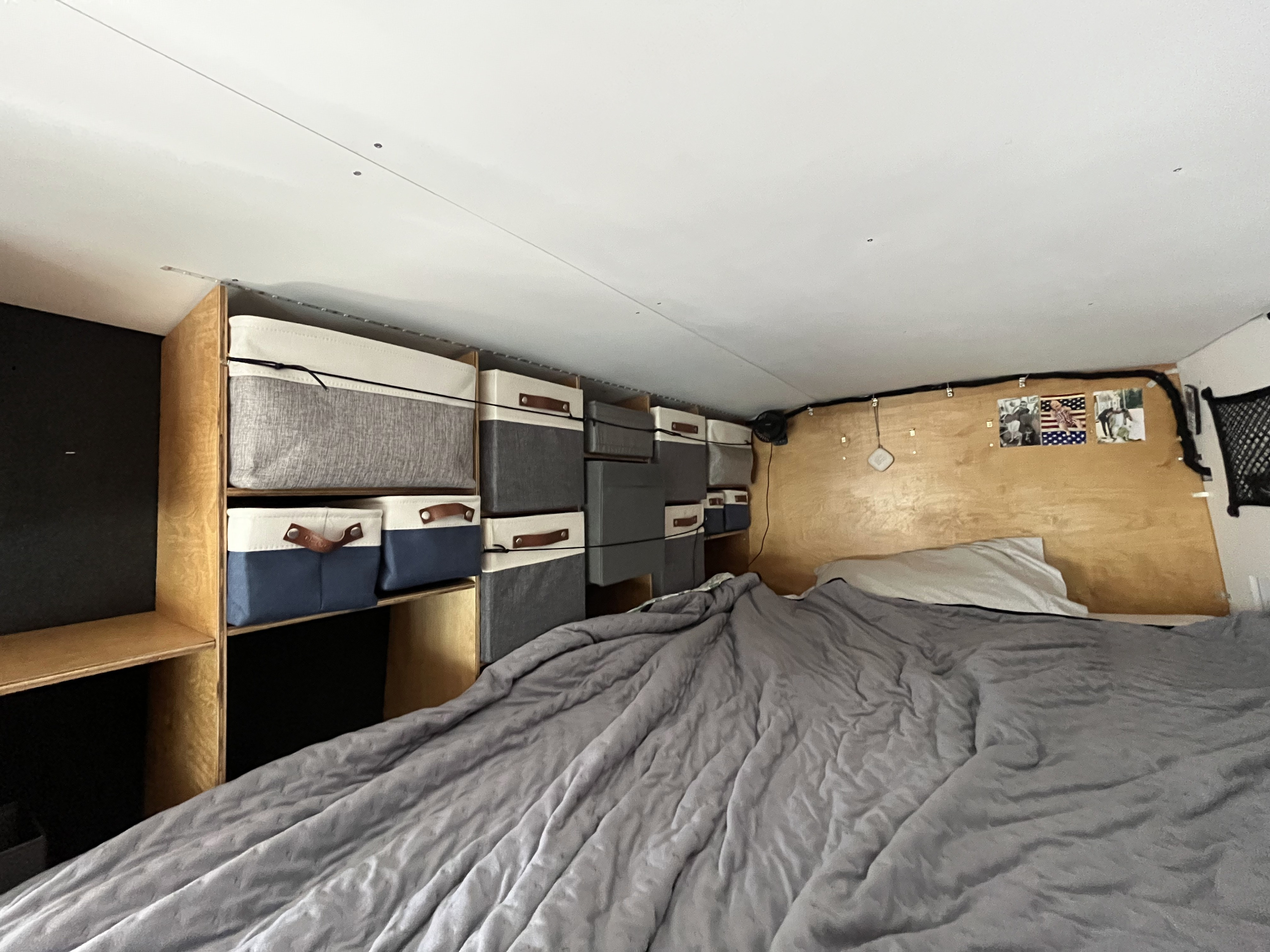 RV rental interior with a cozy bed, gray comforter, and efficient overhead storage in fabric bins on wooden shelves.. Ford Transit 2015