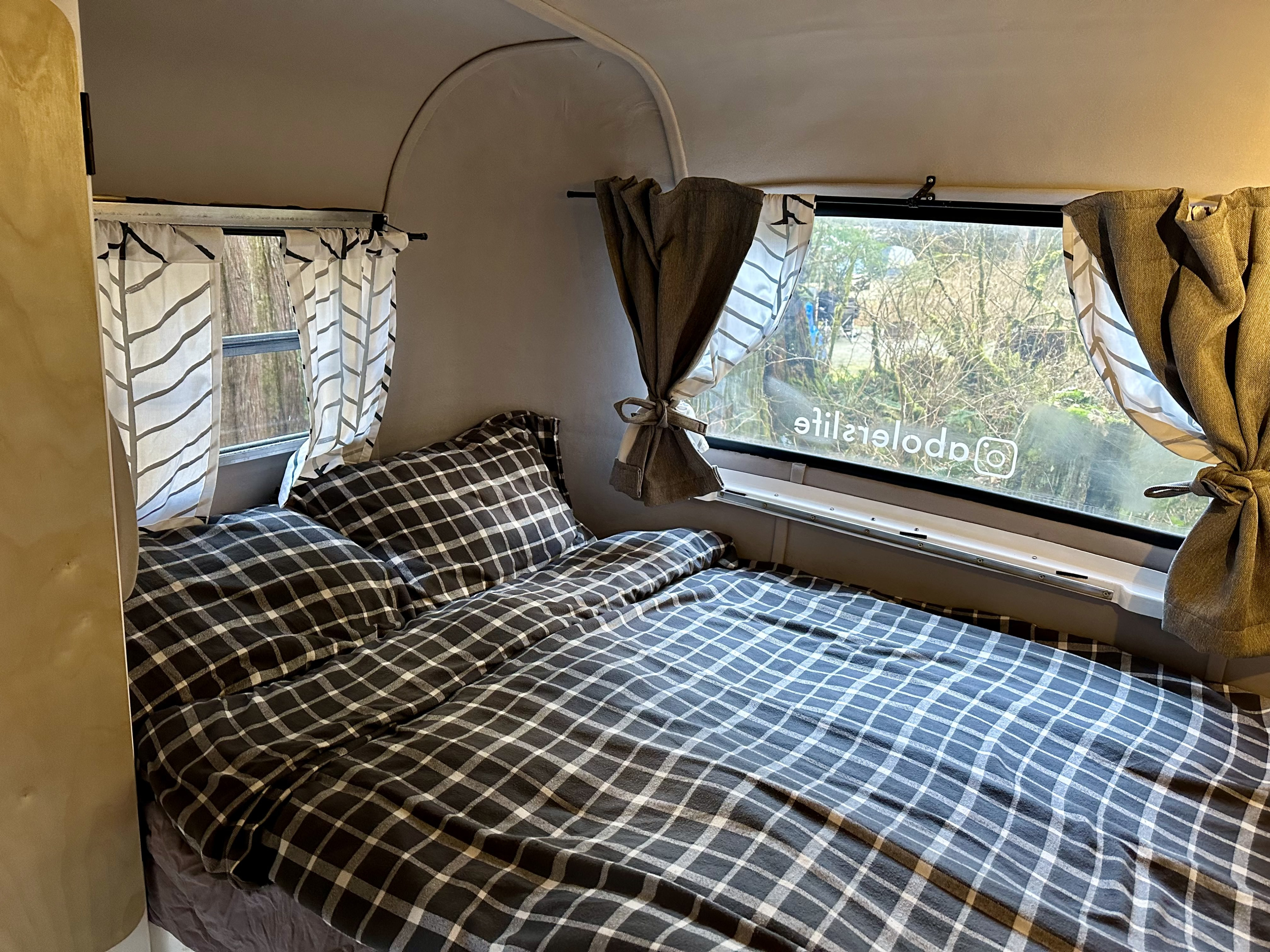 Relaxing RV rental bedroom features a comfy plaid bed, pillows, and windows with curtains overlooking nature.. Boler 1978 Boler 1978