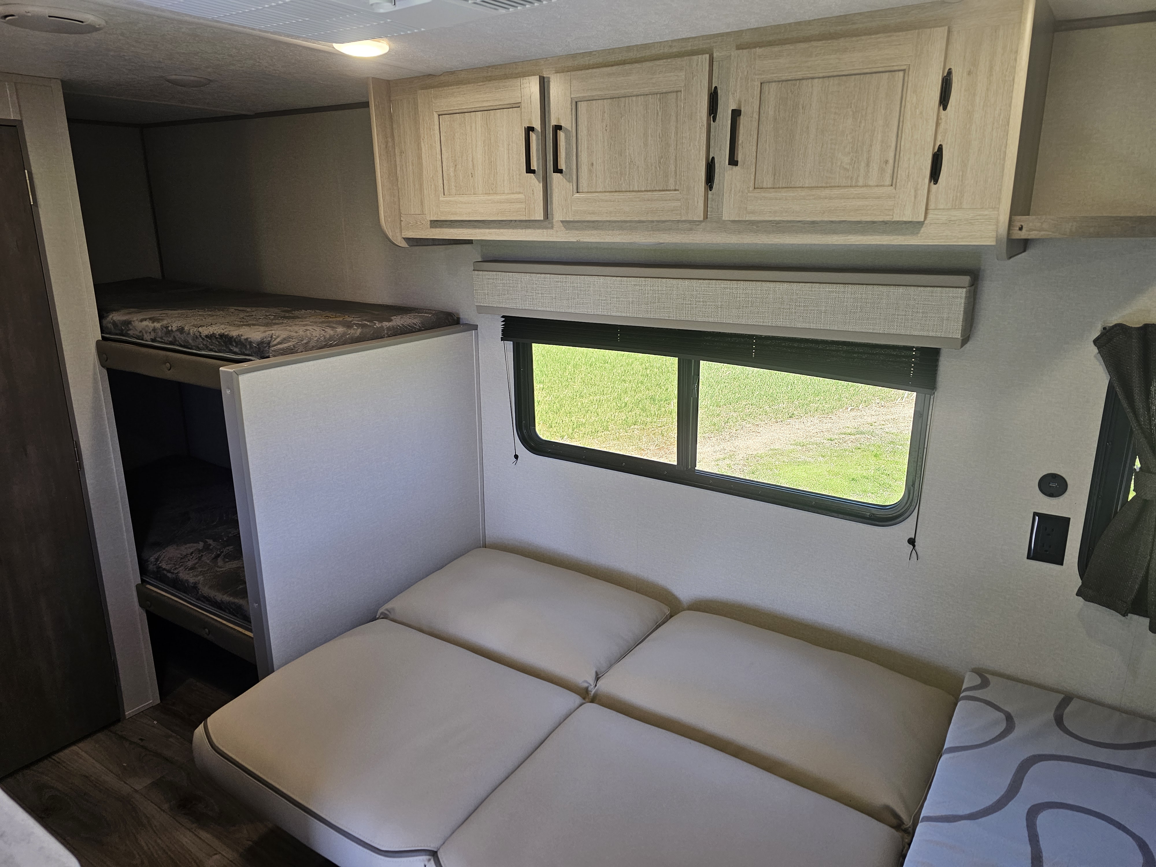 RV rental interior: bunk beds on left, light wood cabinets above, and a convertible sofa by a window to grass.. Forest River Catalina 16BHX 2024