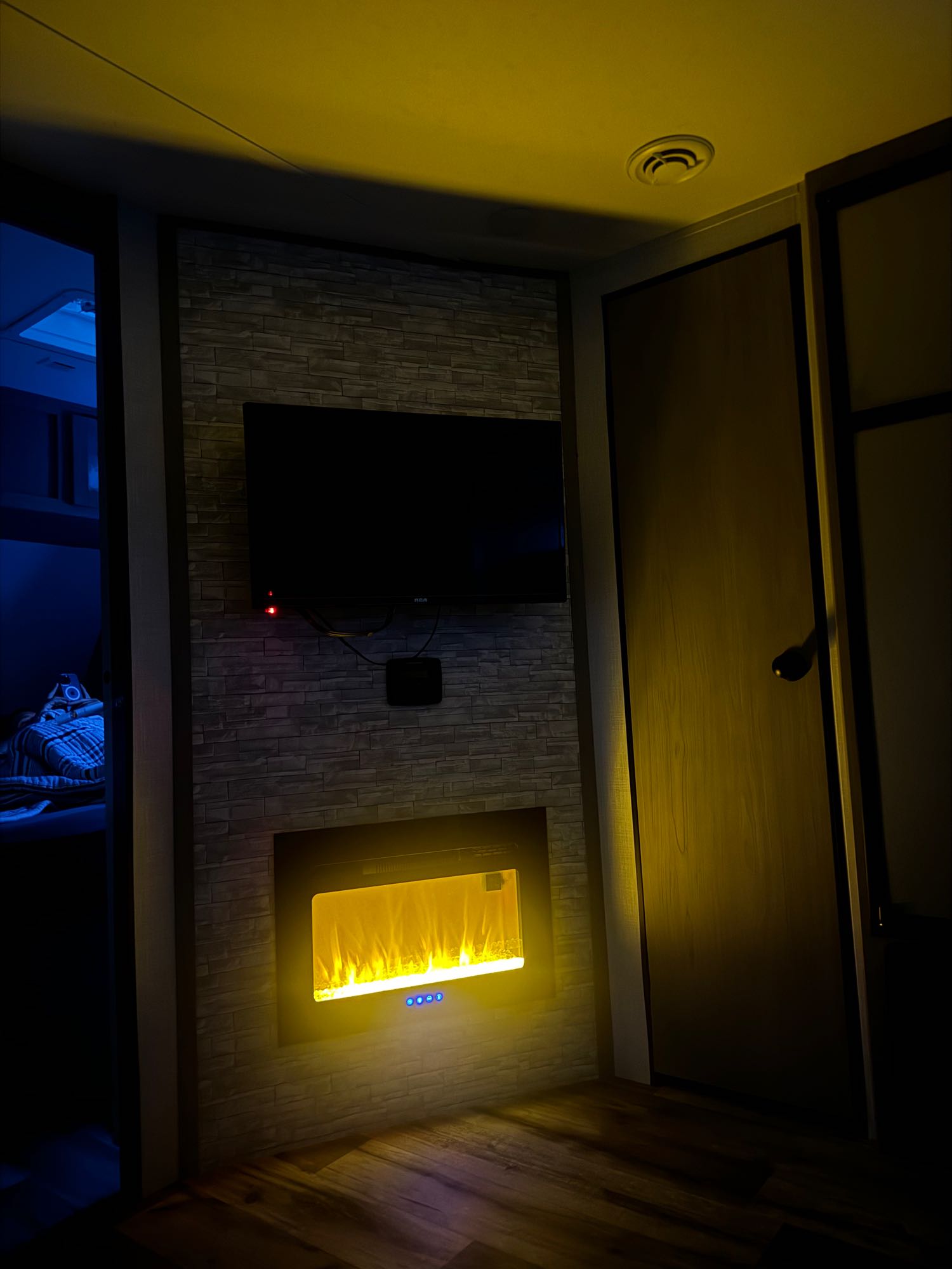 Inviting rv rental interior: TV over a brick-textured wall with glowing electric fireplace. Blue light from adjacent room.. Dutchmen Coleman Light 2023