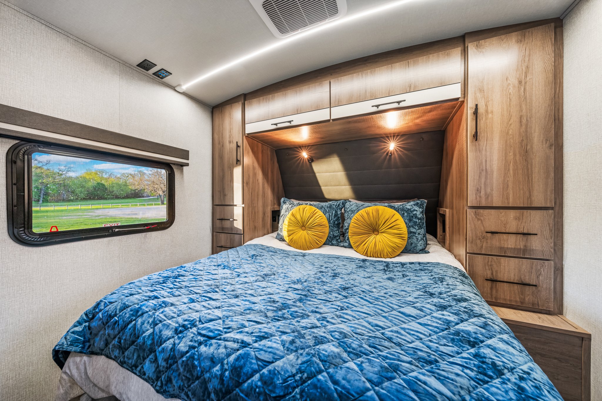 A well-appointed RV rental bedroom with a quilted blue bed, storage, and a window view, ideal for travel comfort.. Grand Design Imagine 2024