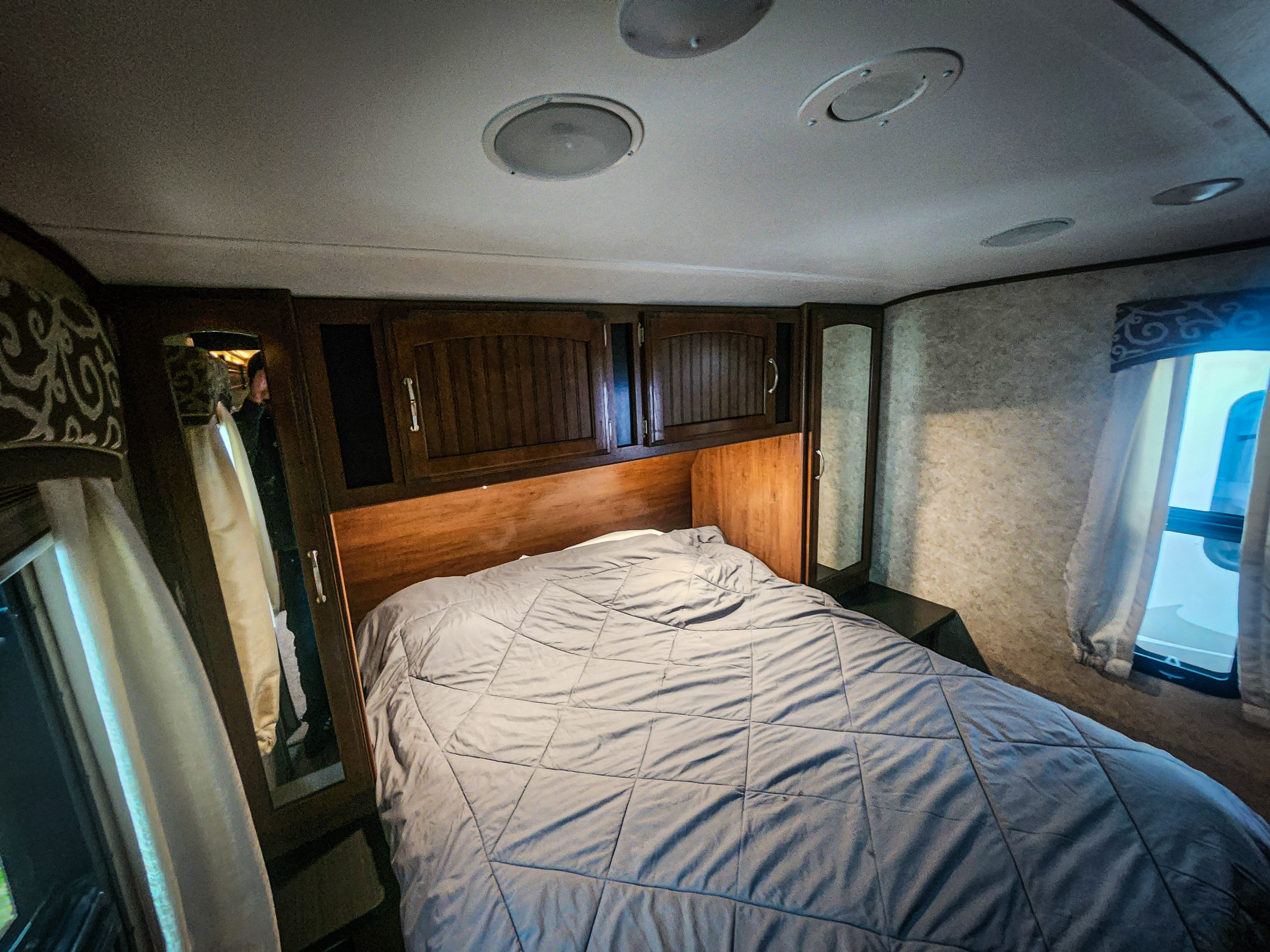 Inside an RV rental bedroom showing a large bed, wooden cabinets, mirrors, and windows. A reflection is seen in the left mirror.. Jayco White Hawk 2017