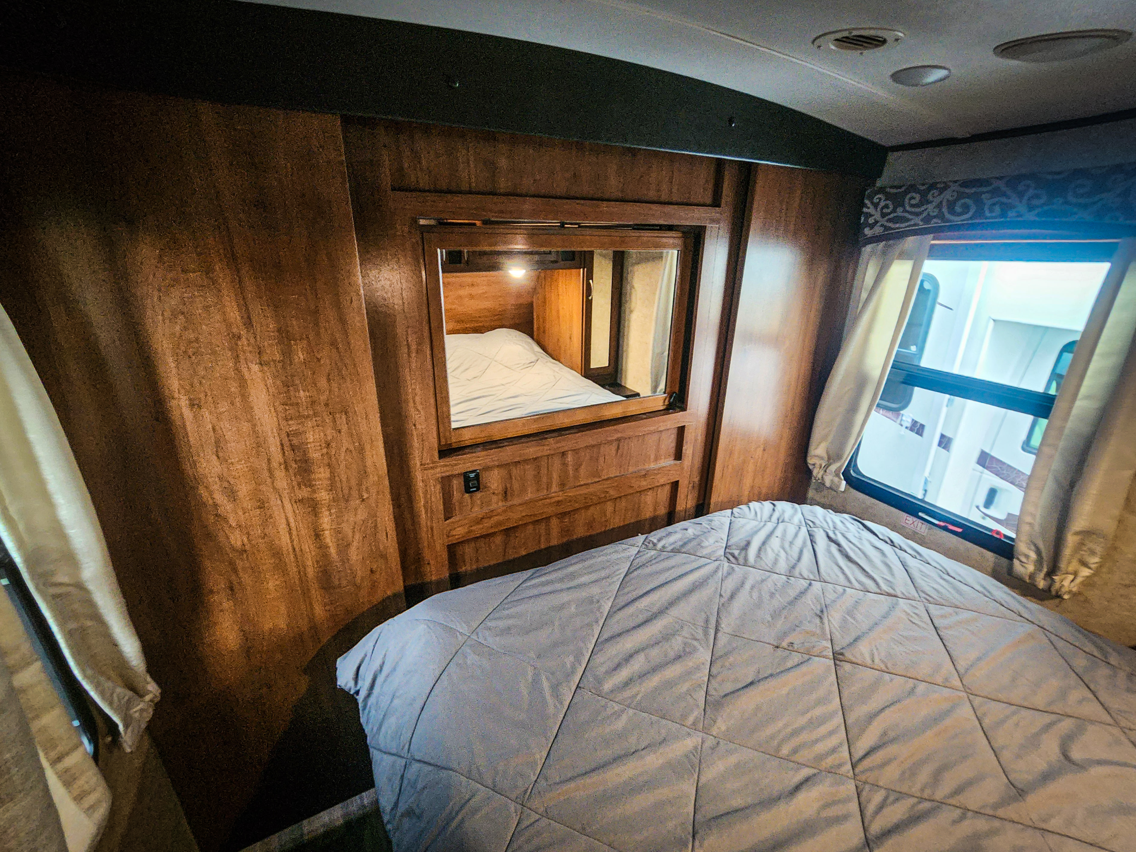 Cozy RV rental bedroom featuring a comfy grey bed, warm wooden interior, and a clear window view for your travels.. Jayco White Hawk 2017