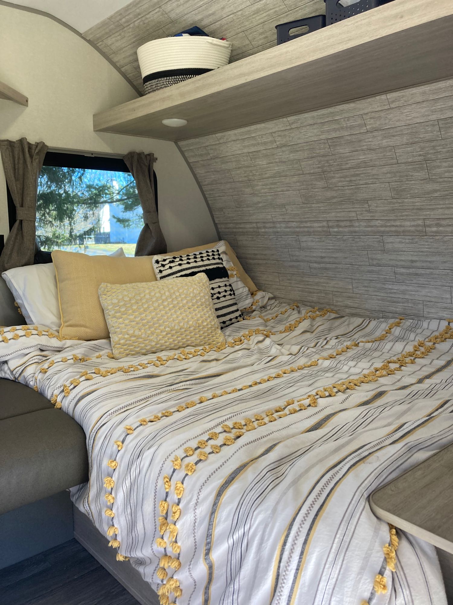 This cozy RV rental features a bed with yellow and striped bedding, decorative pillows, a window with curtains, and overhead storage.. Forest River Aurora Aurora Light 2024