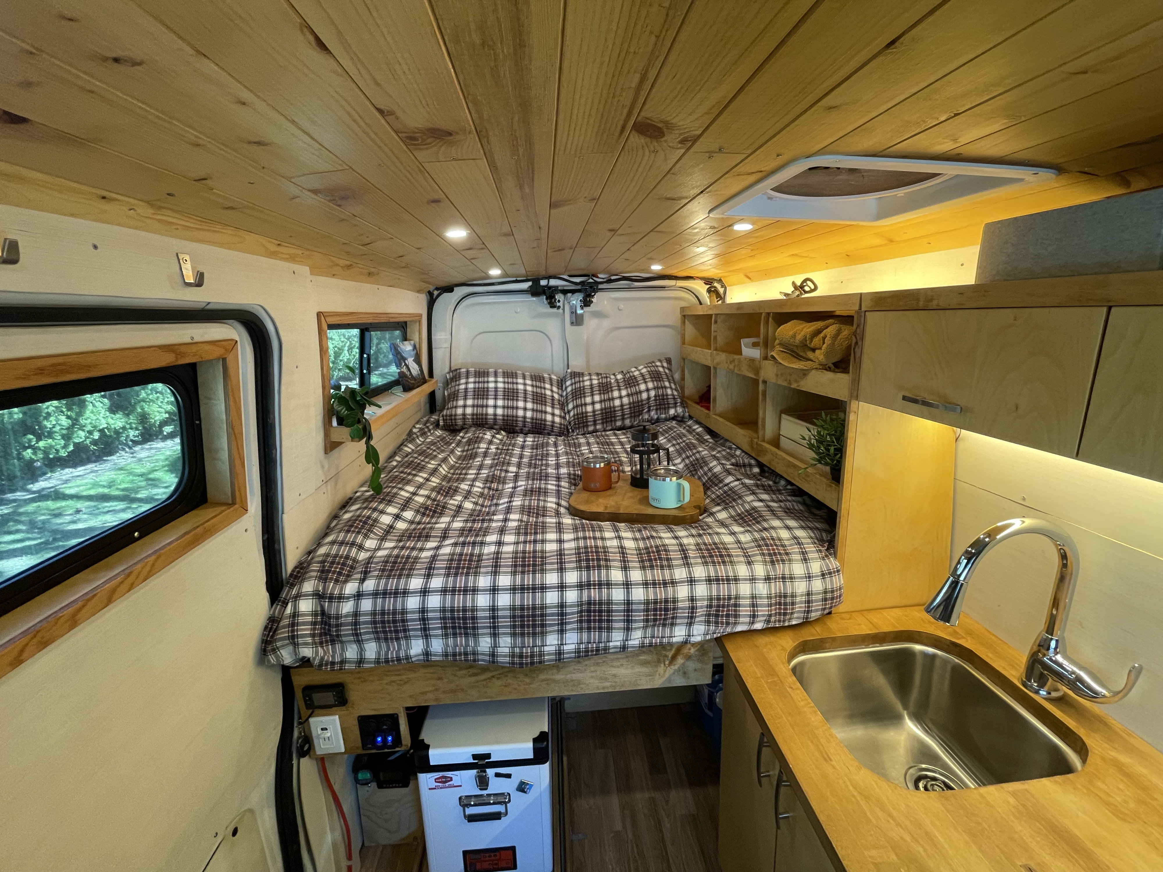 Cozy RV rental interior featuring a large bed with plaid bedding, compact kitchen, and warm wooden paneling.. Nissan NV2500 Cargo Van 2012