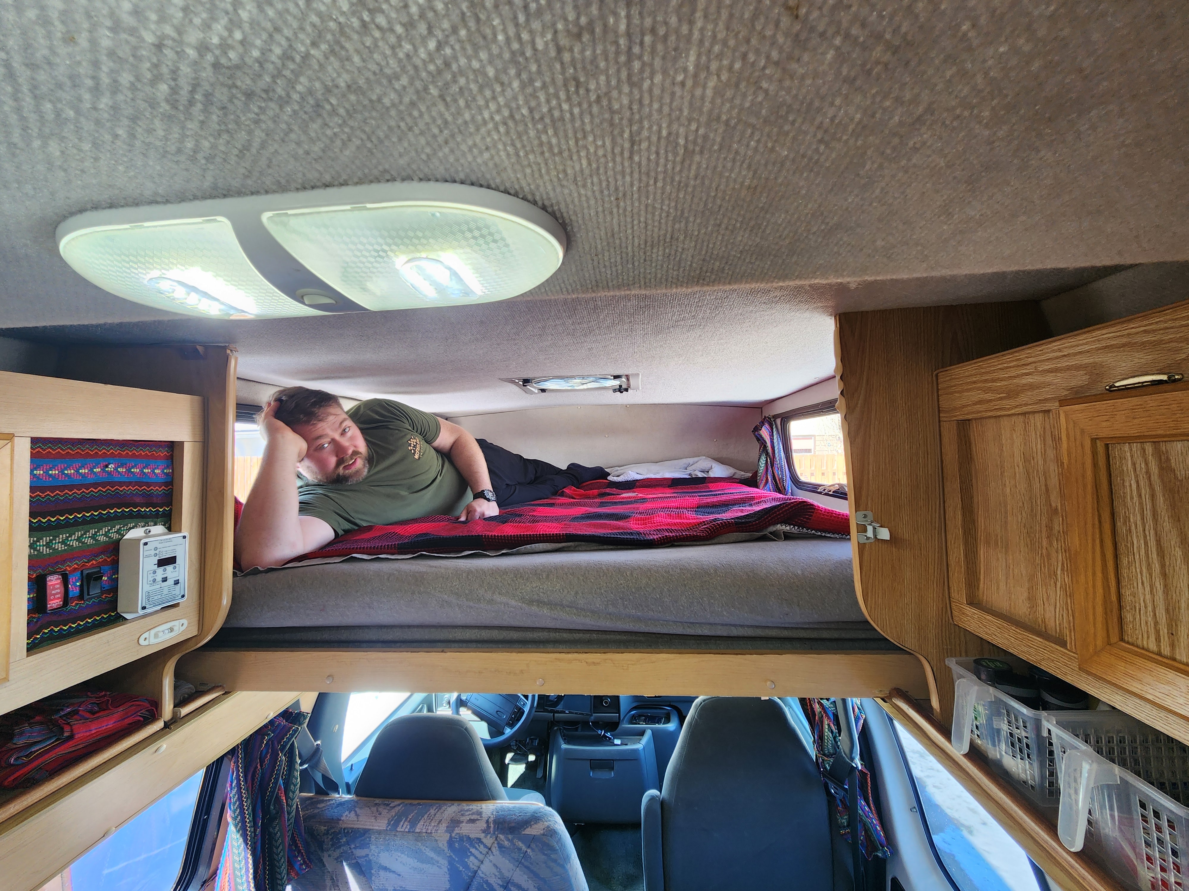 Man relaxing on cozy plaid bunk bed in an RV rental. View shows comfortable interior & front cabin for travel.. Okanagan Adventure Series 1996