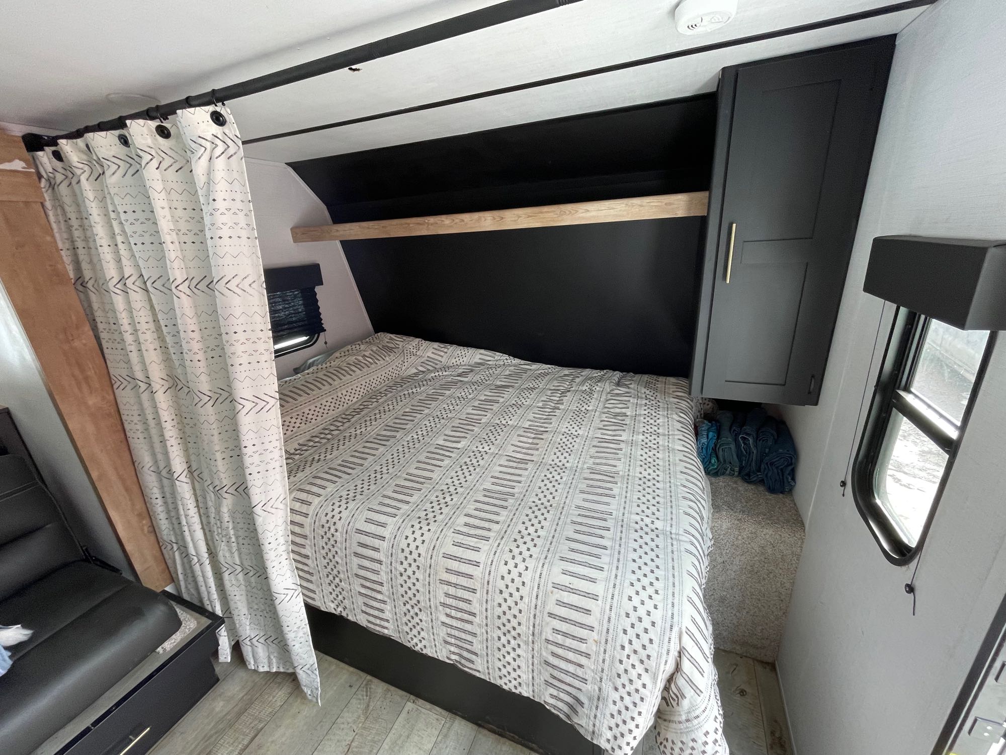 Modern RV rental bedroom with patterned bed, black headboard, wood shelf, and privacy curtain. Compact and stylish RV interior.. Keystone RV Springdale 2023