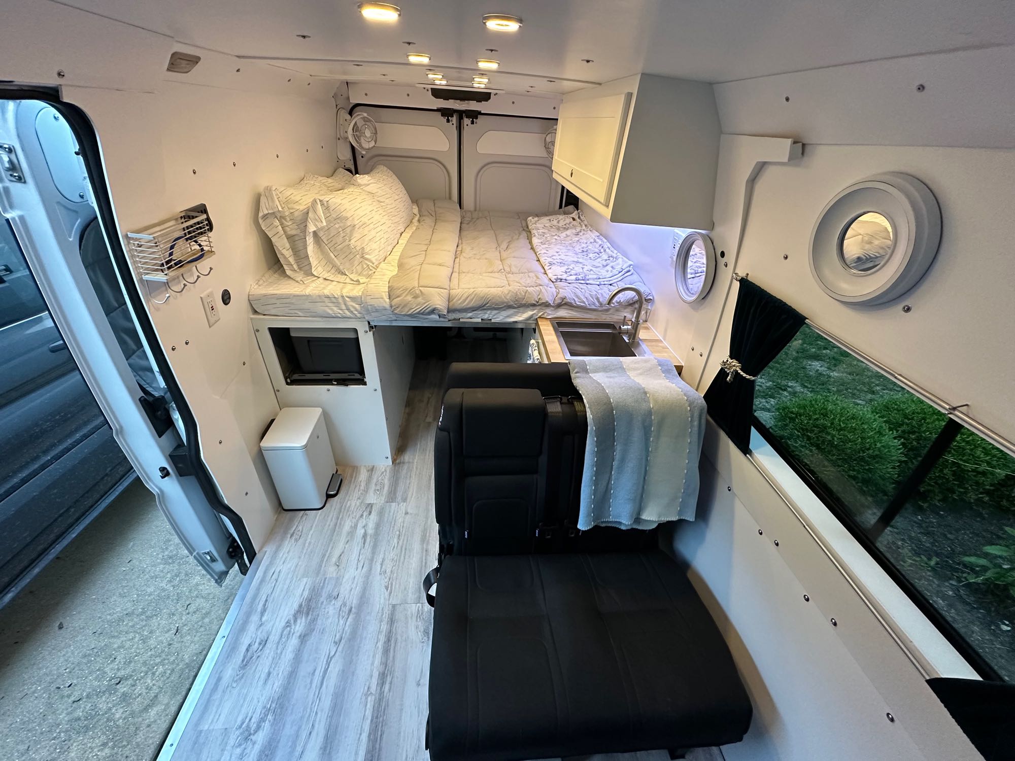 Spacious custom RV rental interior features a cozy bed, compact kitchen, and convertible seating with unique porthole windows.. RAM PROMASTER 2500 2023