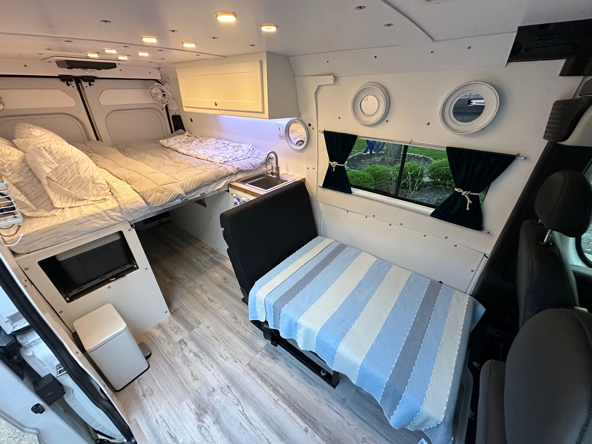Inside an RV rental van: a cozy bed, compact kitchenette, and passenger seating area. Wood-look floors and ample lighting create a comfortable living space.. RAM PROMASTER 2500 2023