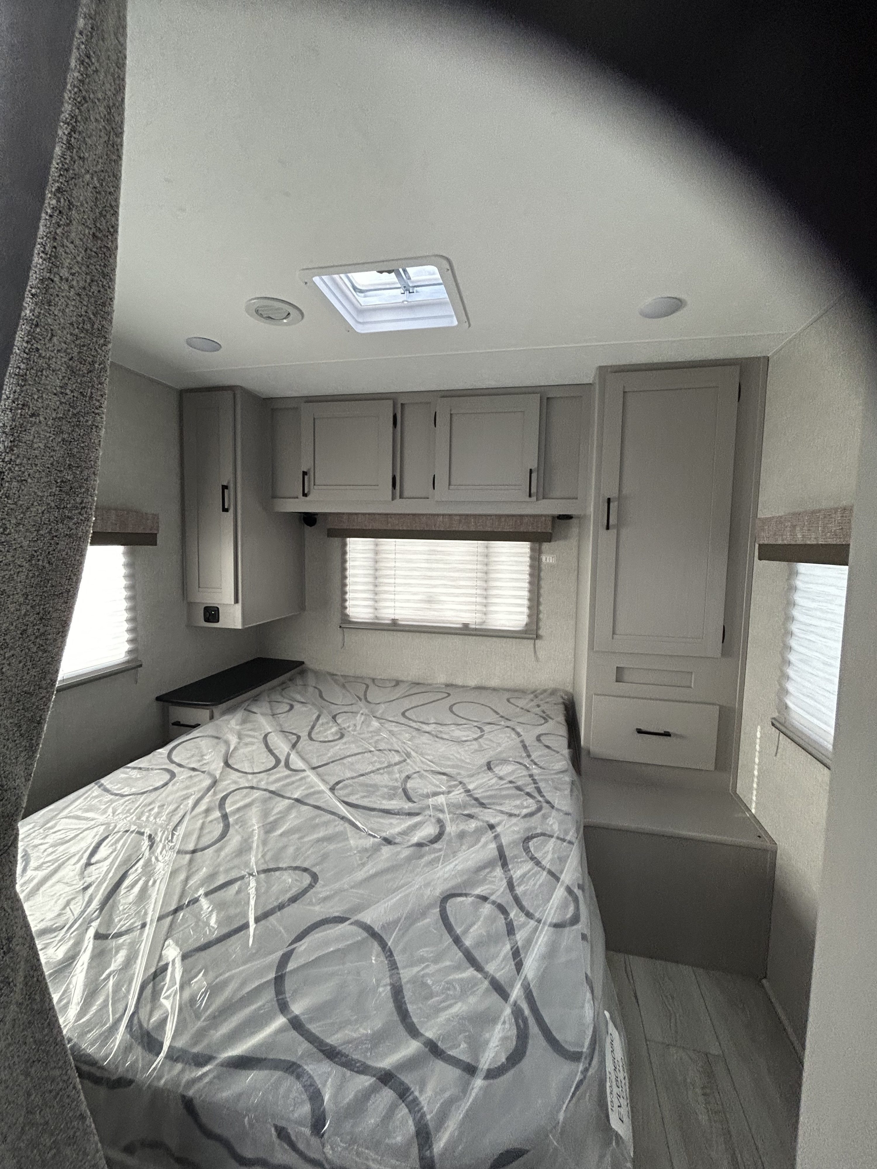 RV rental interior: A bright, neutral-toned bedroom with a plastic-covered bed, overhead cabinets, and window blinds.. Forest River Freelander 2024