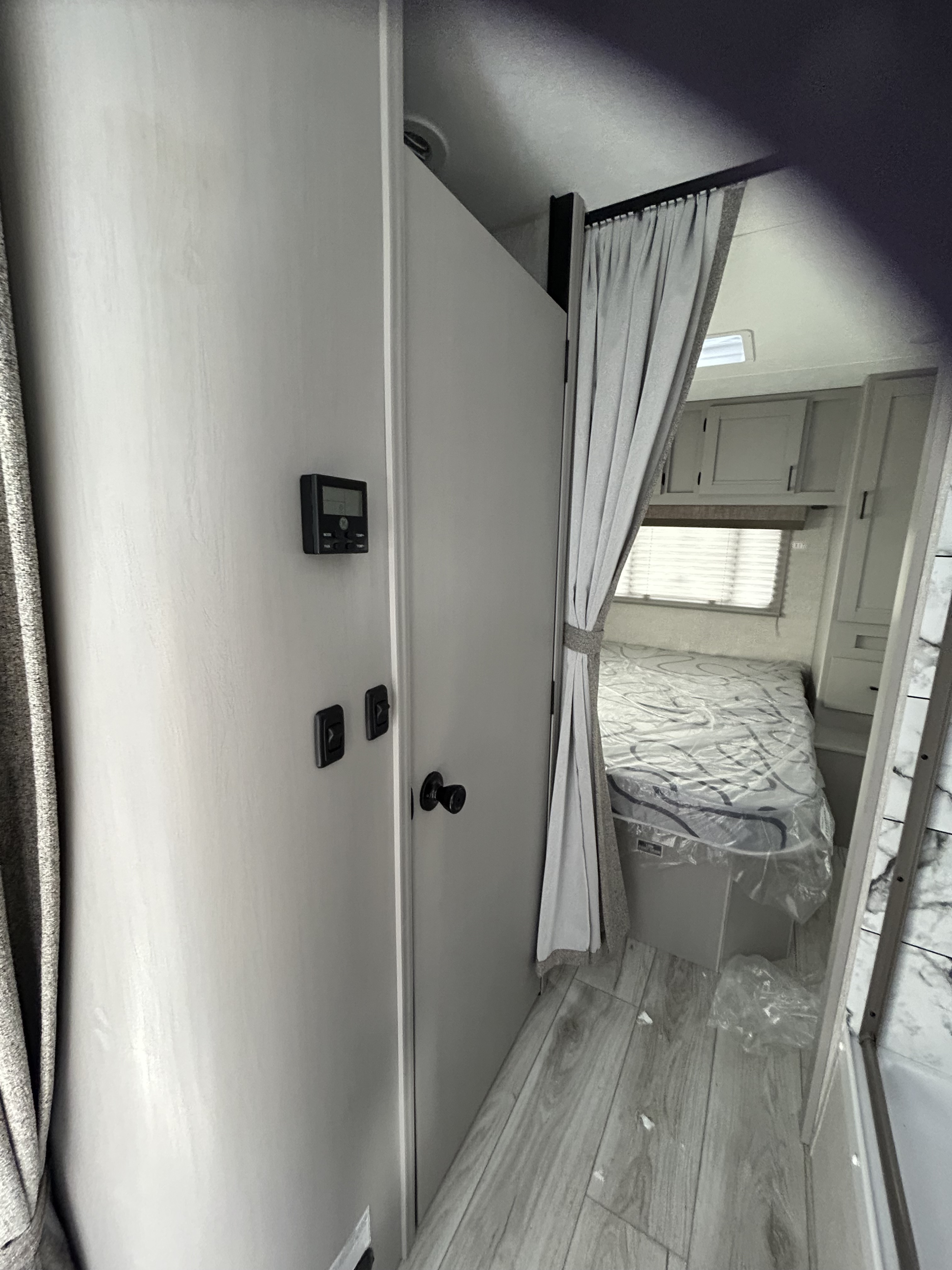 RV rental interior: Bedroom with a bed (plastic-covered), window, cabinets, and wall controls. Light-colored walls and wood-look floor.. Forest River Freelander 2024