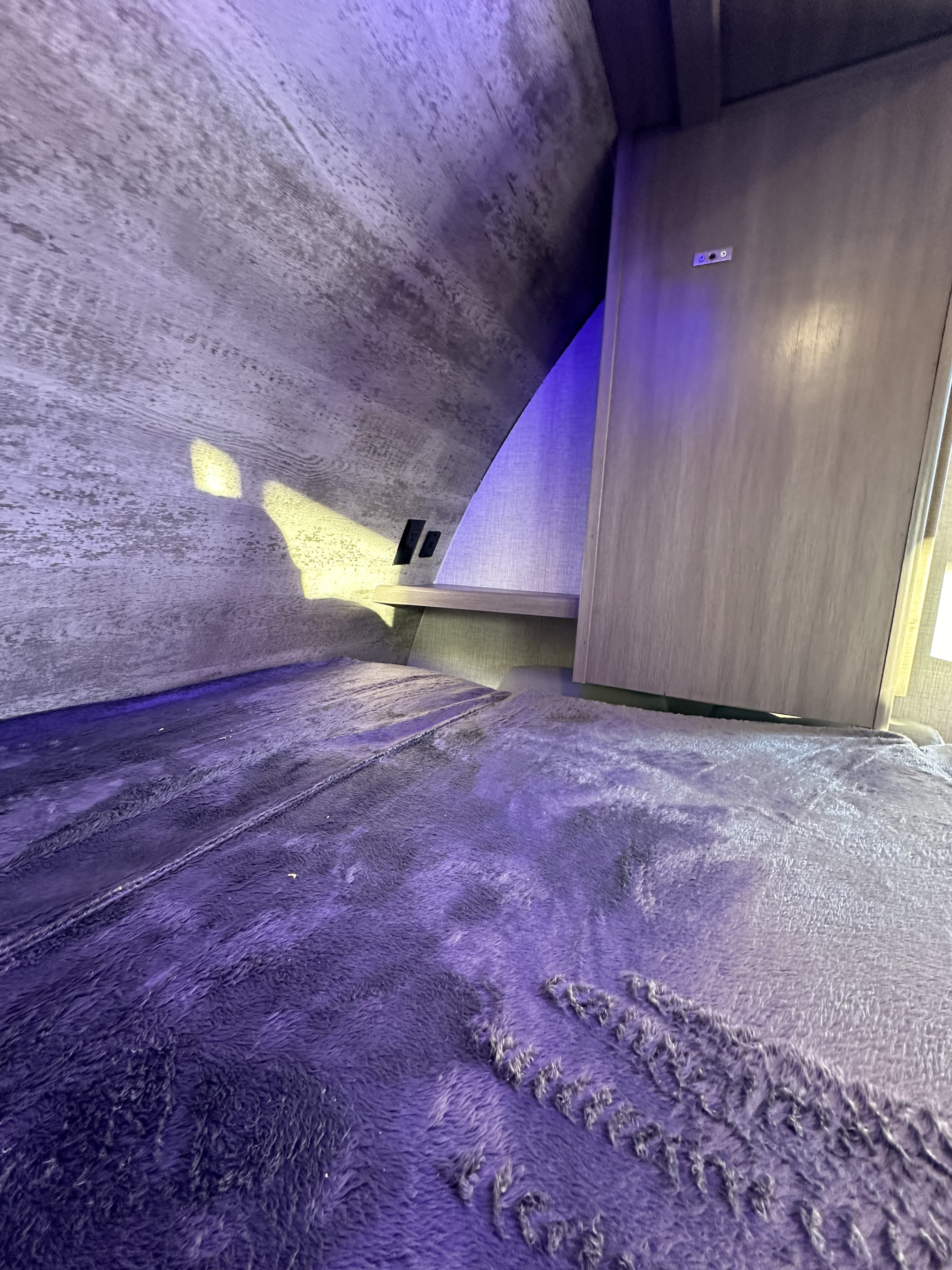 Inside an RV rental bunk: a soft purple bed, wood-paneled walls, a small shelf with outlets, and ambient lighting.. Crossroads RV Sunset Trail Super Lite 2022