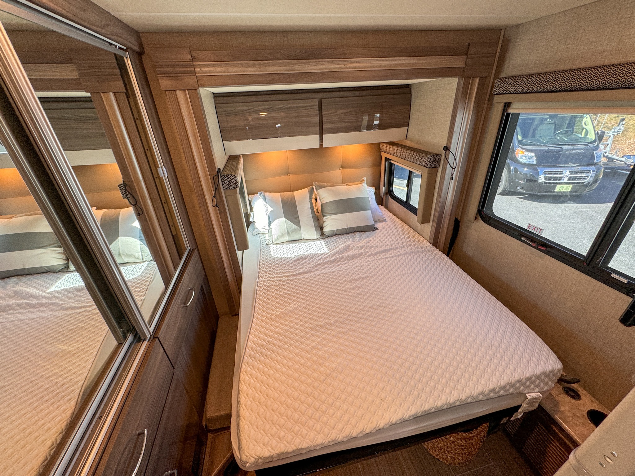 Inside a modern RV rental: a cozy bedroom with a quilted bed, overhead cabinets, and a window view to the outdoors.. Thor Motor Coach Delano 2022