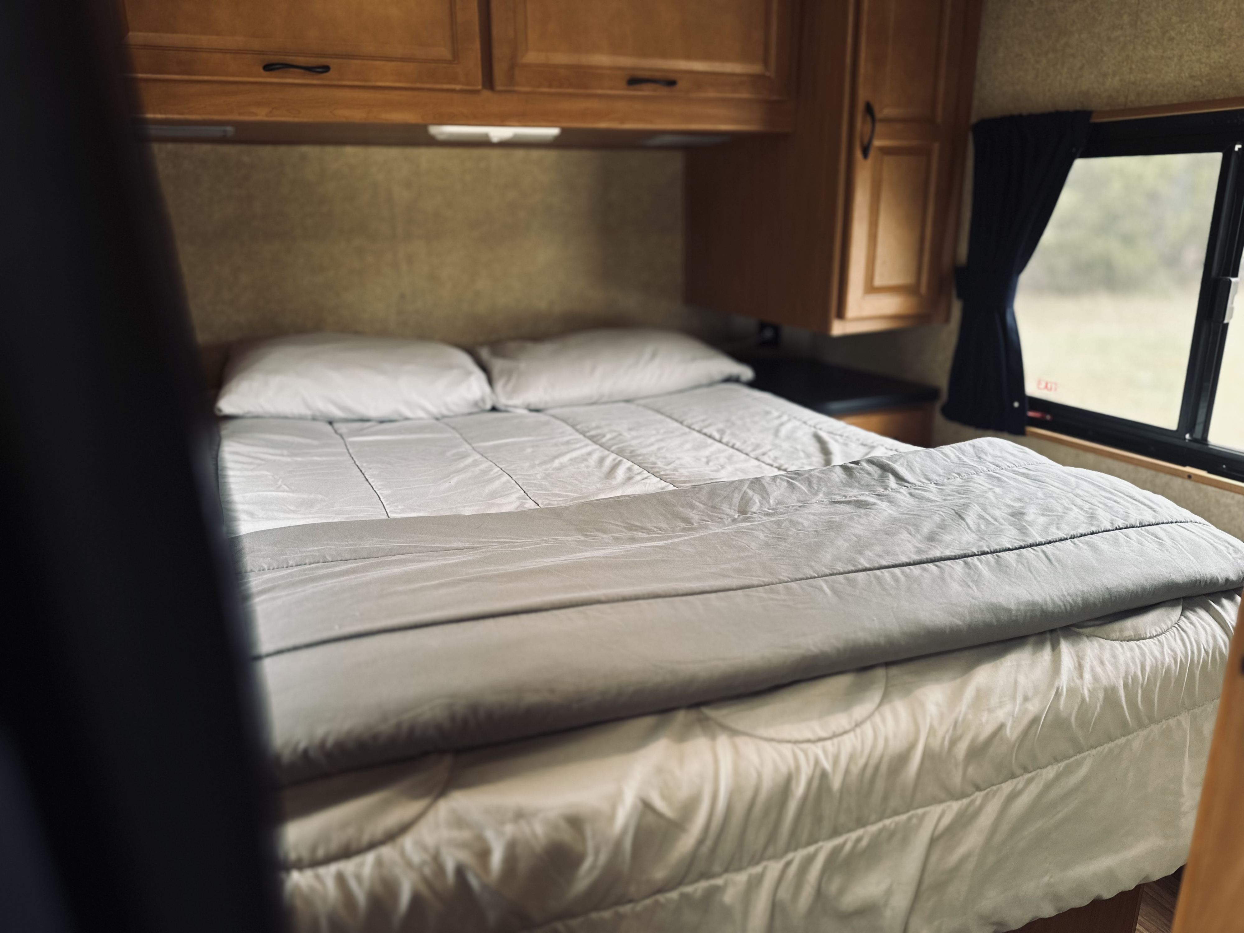 Cozy interior of an RV rental bedroom with a neatly made bed, white and gray bedding, wooden cabinets, and a window.. Thor Majestic 2016