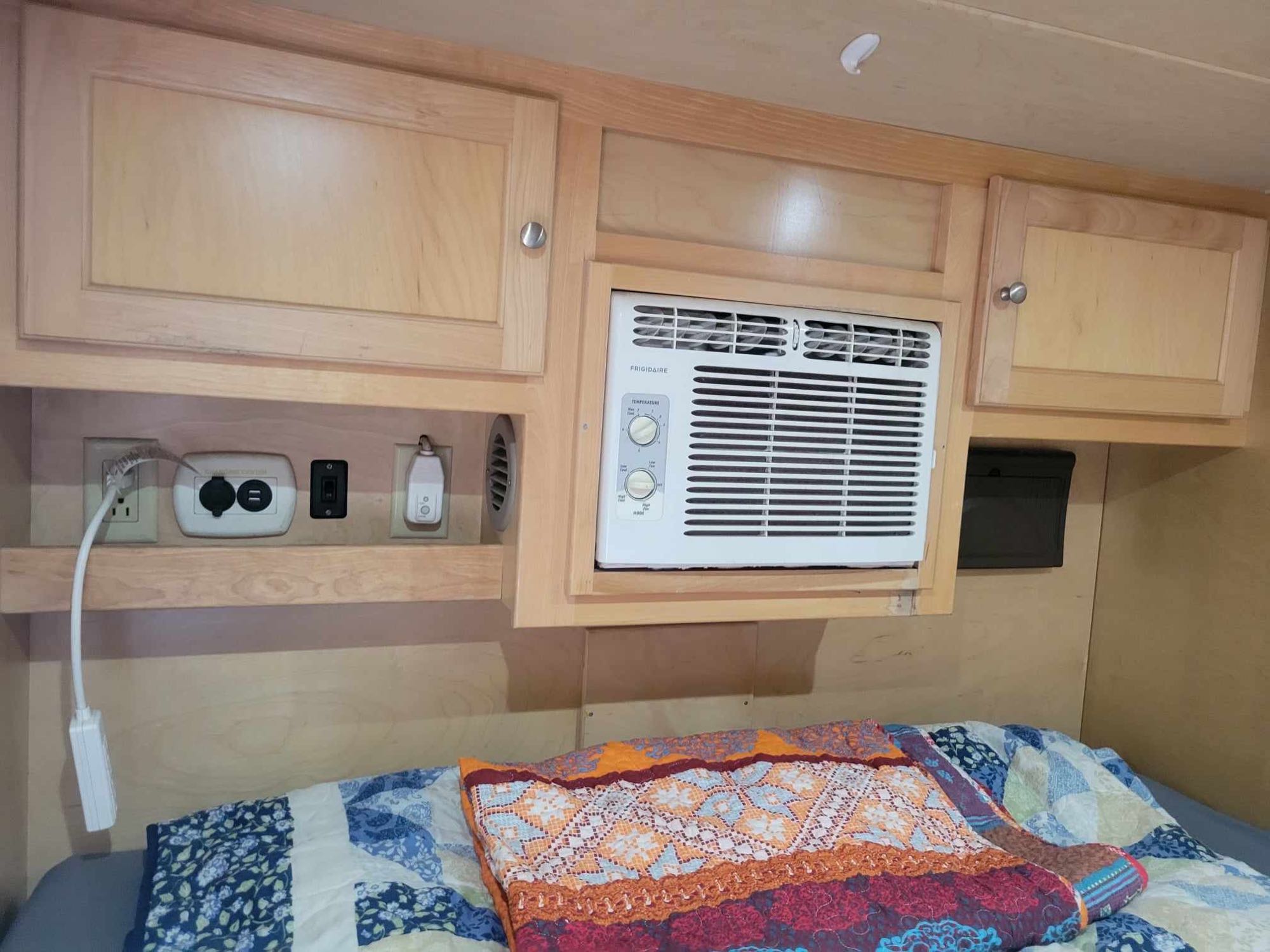 RV rental bedroom: a cozy bed below wooden cabinets, AC, and a shelf with various electrical outlets.. Little Guy Silver Shadow 5x10 2015
