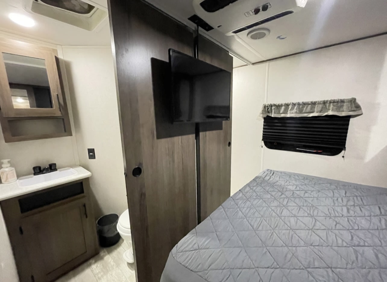 Inside an RV rental: a modern bedroom with quilted bed, wall-mounted TV, and a compact bathroom vanity area.. Forest River PRIME TIME AVENGER 2021