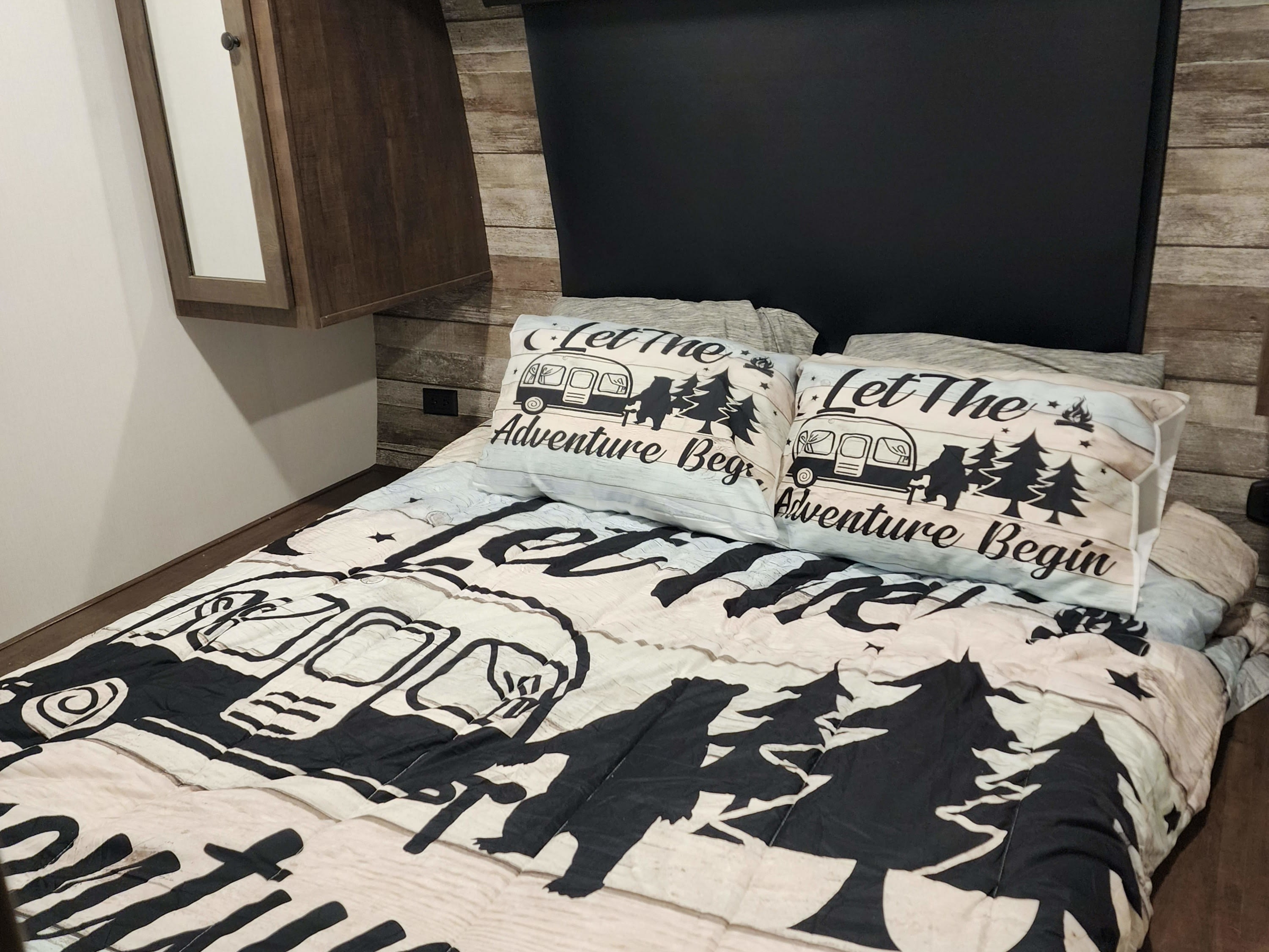 An RV rental bedroom features a cozy bed with adventure-themed bedding, "Let The Adventure Begin" pillows, and wood walls.. Forest River Hyperlite 2815 2022