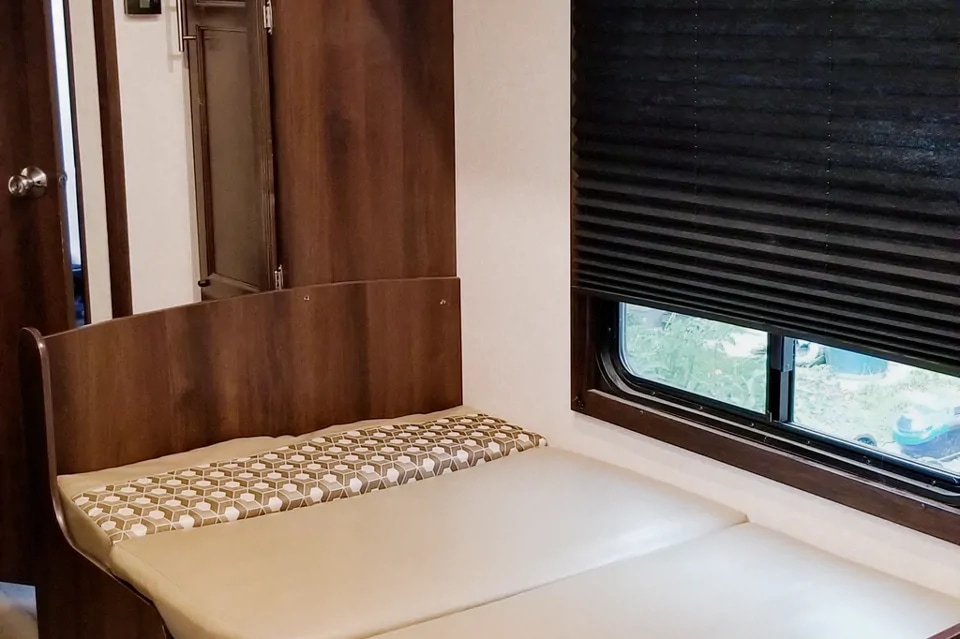 RV rental interior featuring a comfy bed/couch, warm wood paneling, and a window with a dark blind.. Jayco Jay Flight SLX Baja Edition 2019