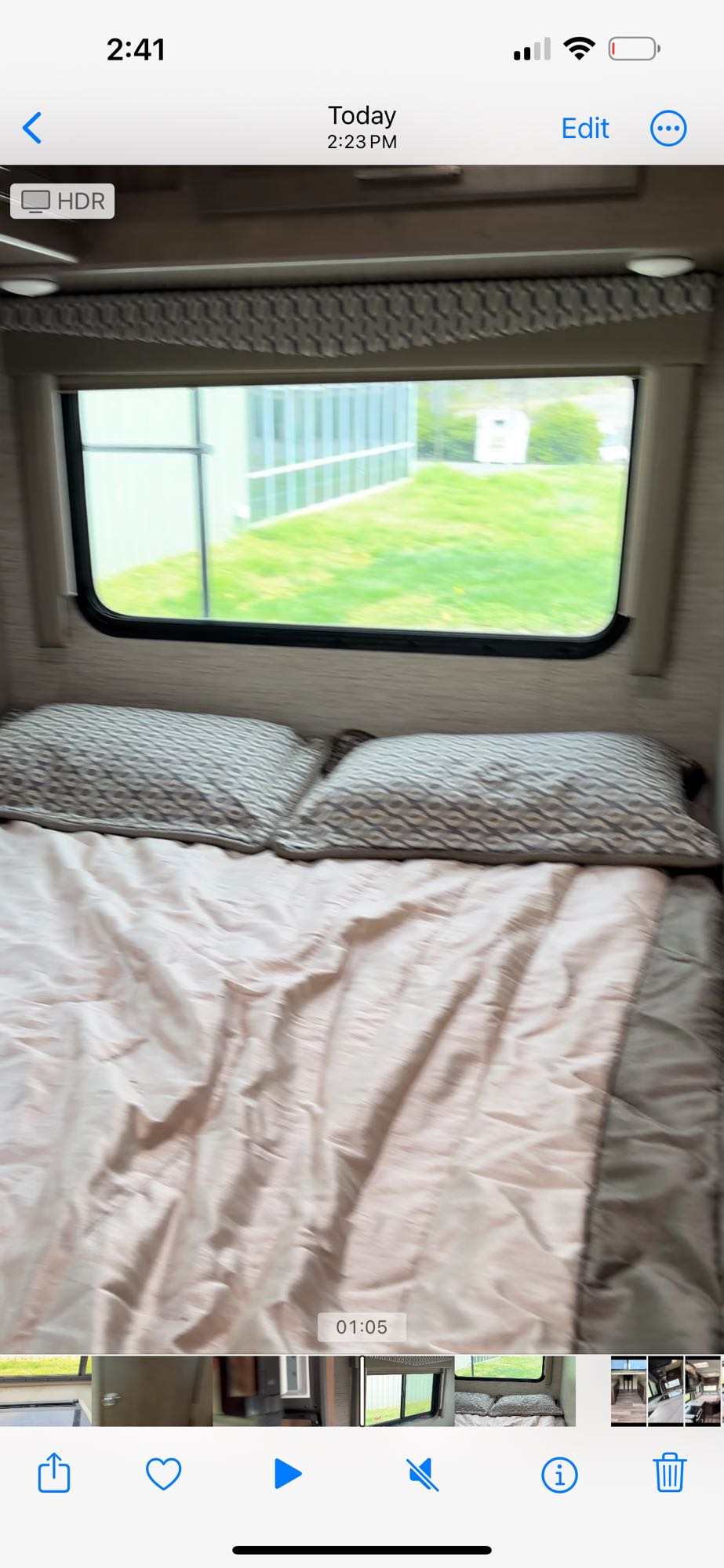 Inside an RV rental: a cozy bed with pillows. The window offers a blurred view of green grass & buildings, hinting at travel.. Thor Freedom Elite 2021