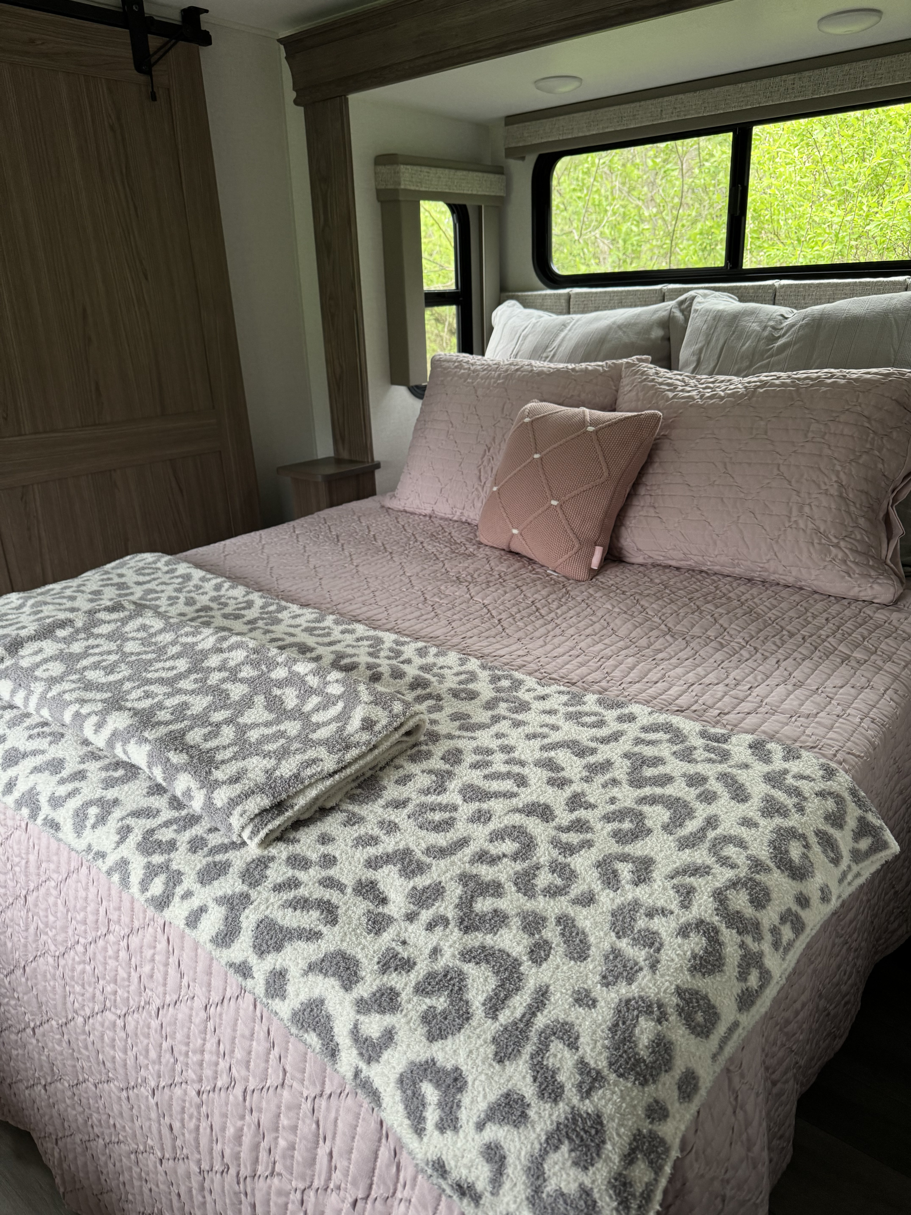 RV rental bedroom: a bed with pink quilted bedding, a leopard throw, and green foliage visible through windows.. Alliance RV Paradigm 2023