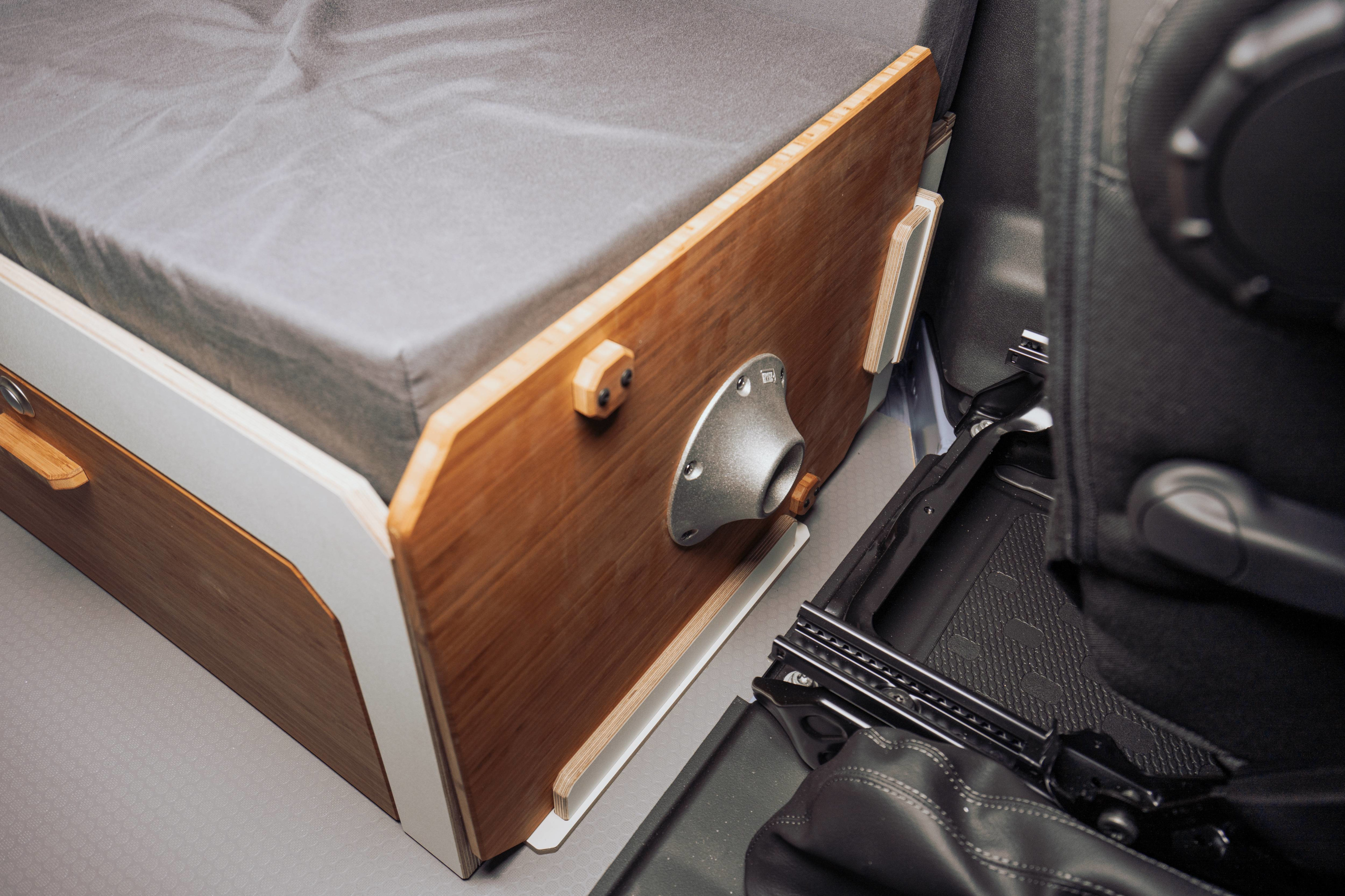 RV rental bed setup: A gray cushioned, modular sleeping and storage unit with wooden panels and a metal fitting inside a vehicle.. Renault Trafic 2025