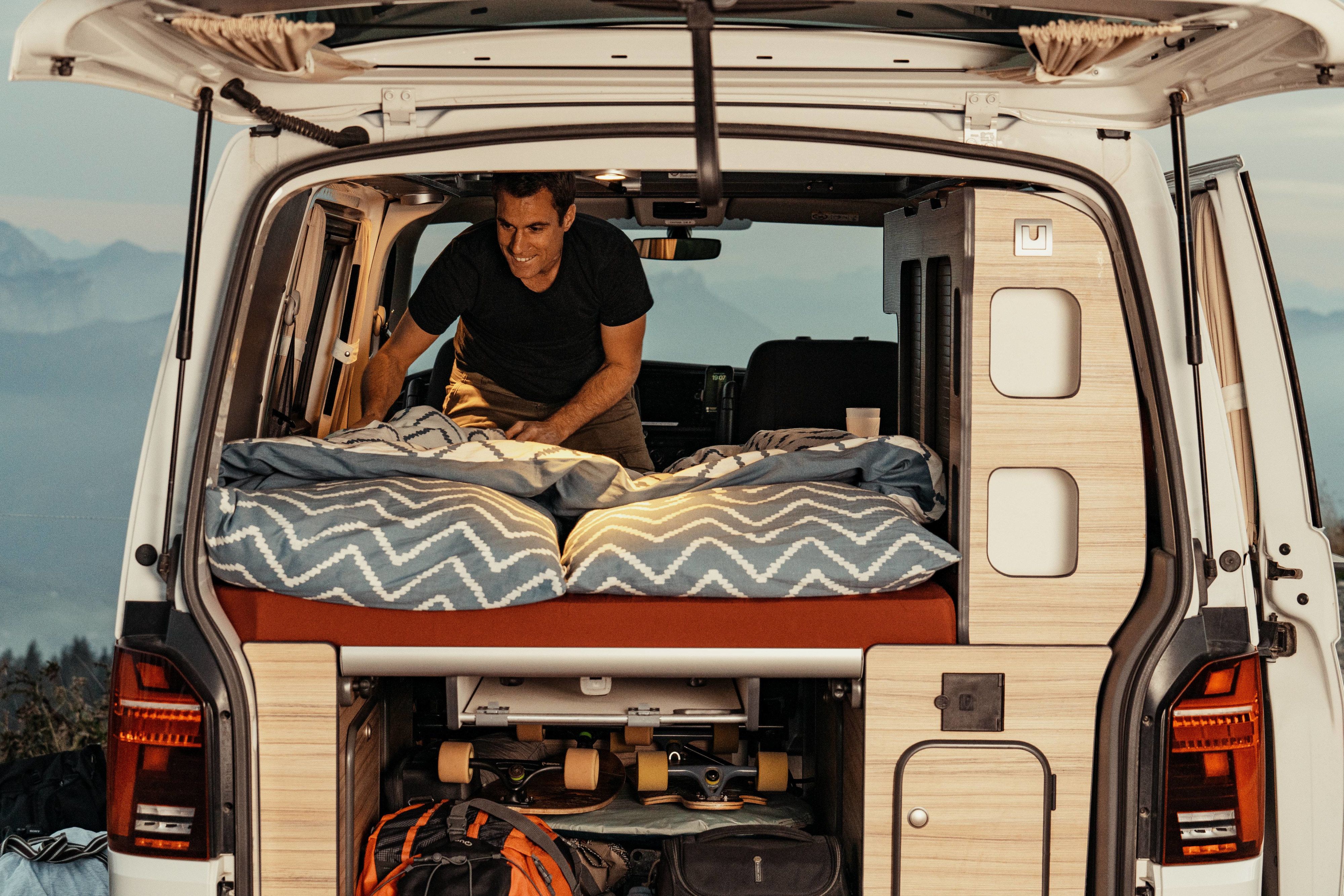 Inside a spacious RV rental, a man smiles while preparing the comfortable bed in a custom camper van, with ample storage below.. Volkswagen Transporter T6.1 2022