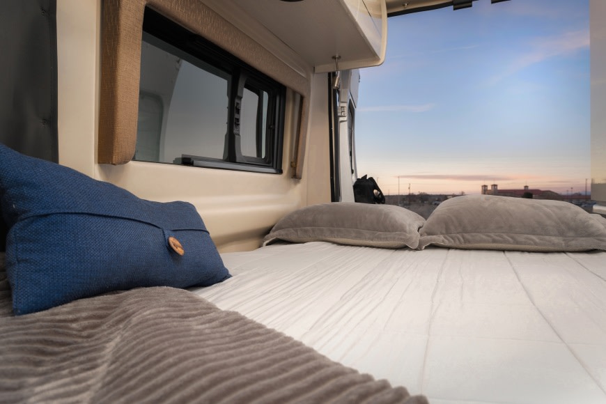 Inside a comfortable RV rental: A soft bed with pillows, offering a peaceful view through the open rear door at dusk.. Thor Rize 2022