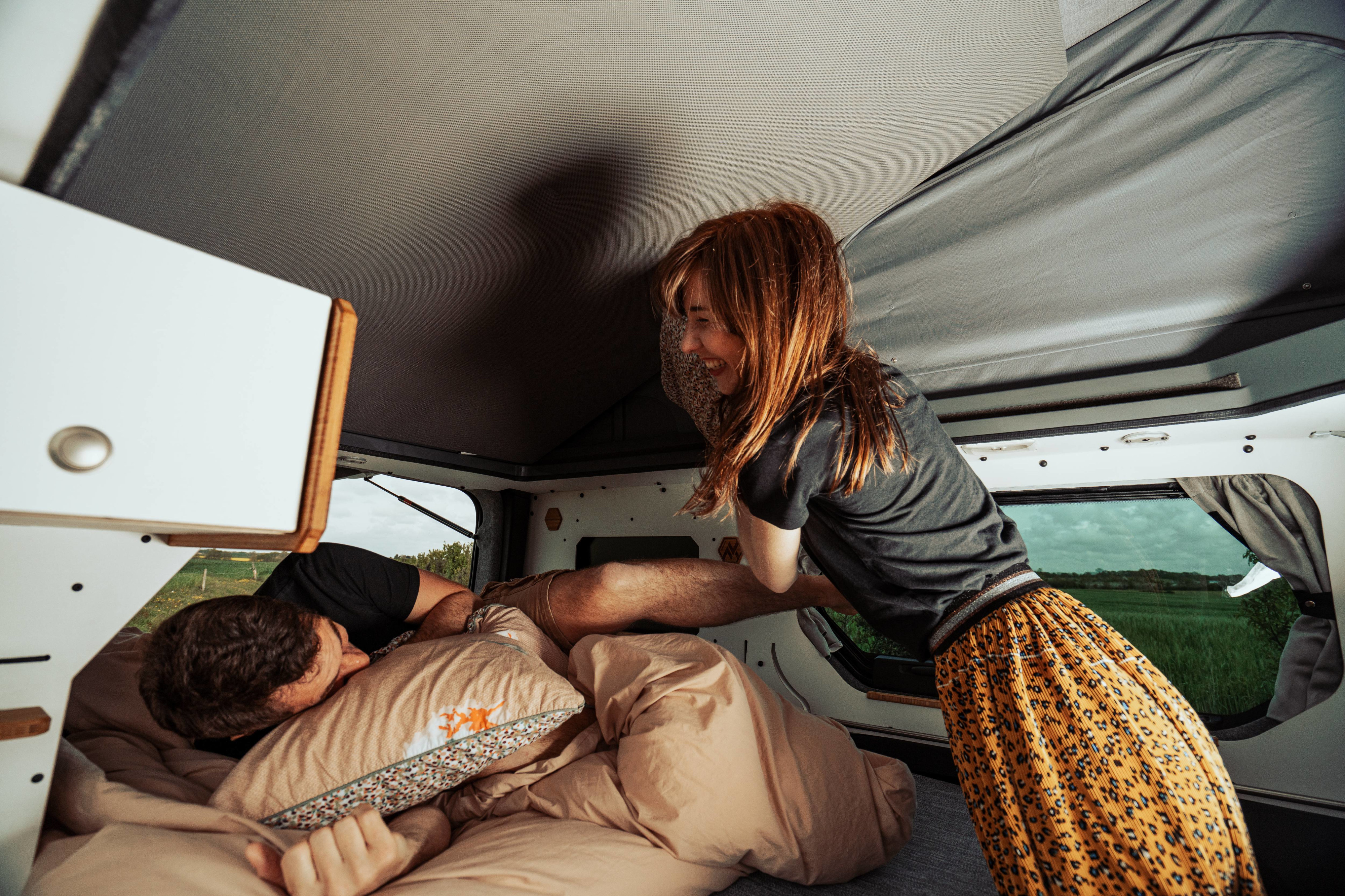Cozy morning in an rv rental! A woman playfully wakes a man sleeping in their camper, surrounded by nature outside.. Renault Trafic 2023