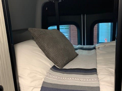 Cozy RV rental bed with white sheets, a grey pillow, and striped blanket. Rear windows show brick. Ready for adventure!. Winnebago Revel 2022