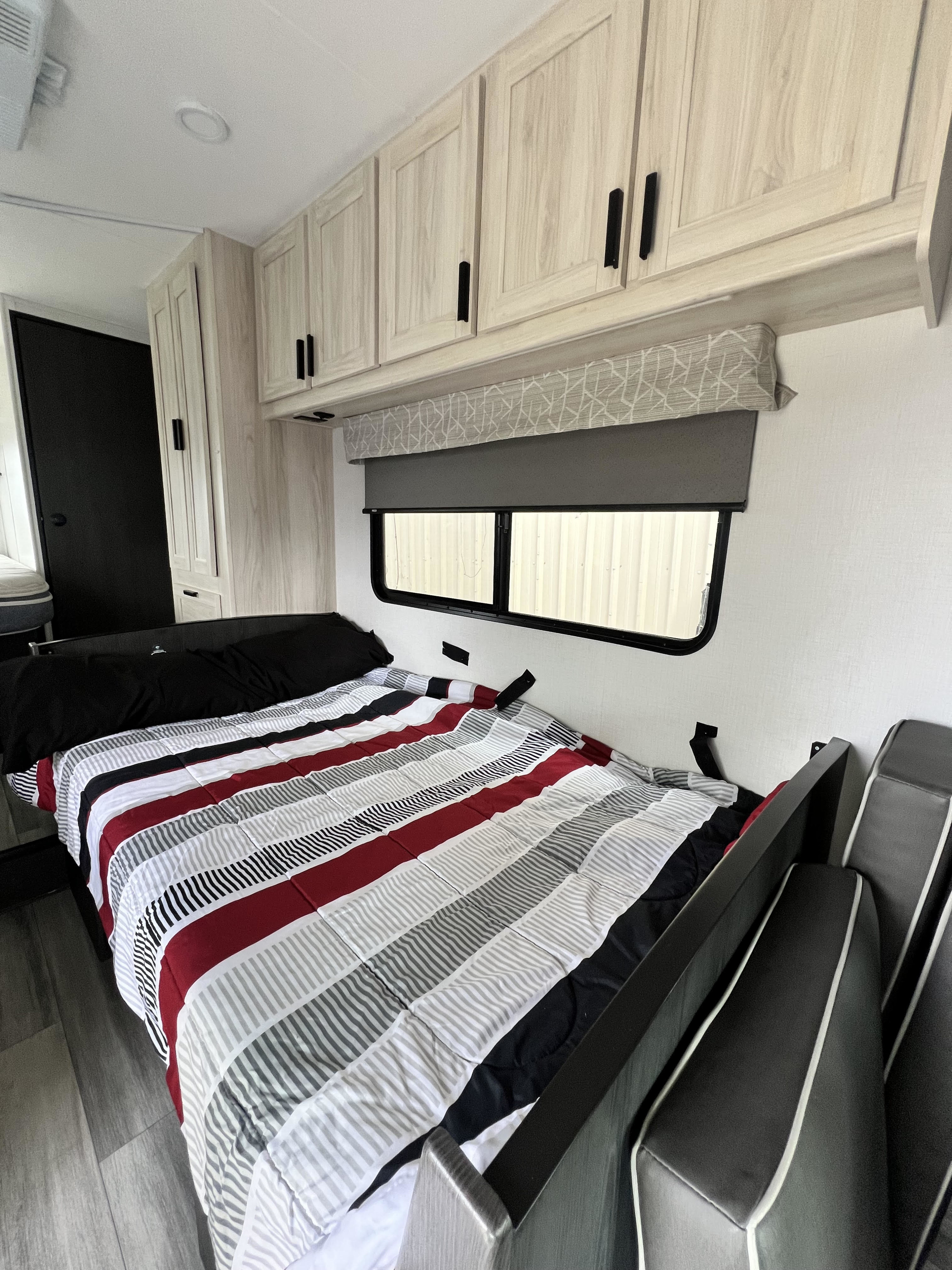 RV rental bedroom with a striped comforter, light cabinets, a window with shades, and dark gray seating.. Forest River Sunseeker 2025